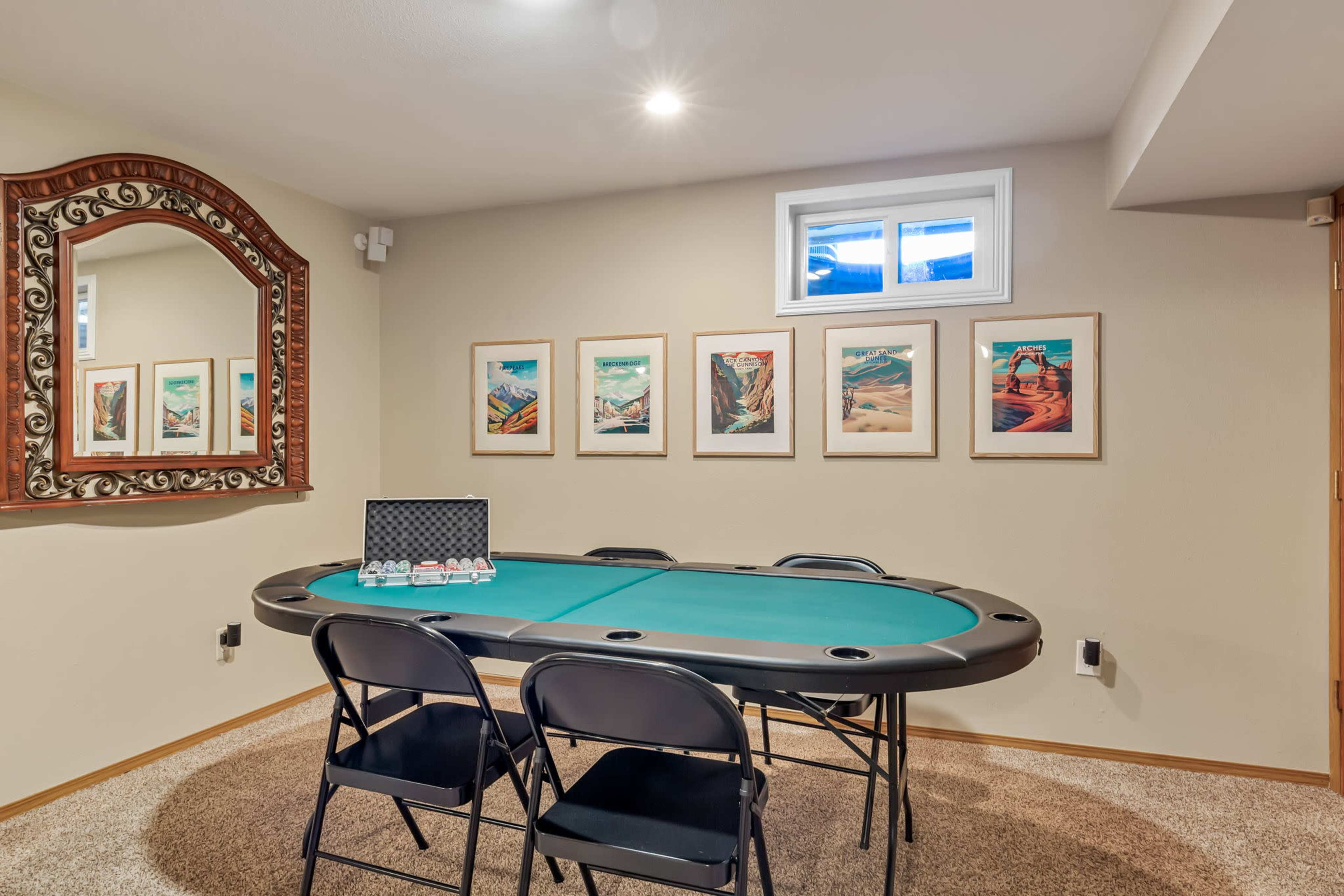 A poker table surrounded by four black chairs is situated in a room with framed artwork on the walls and a large mirror.
