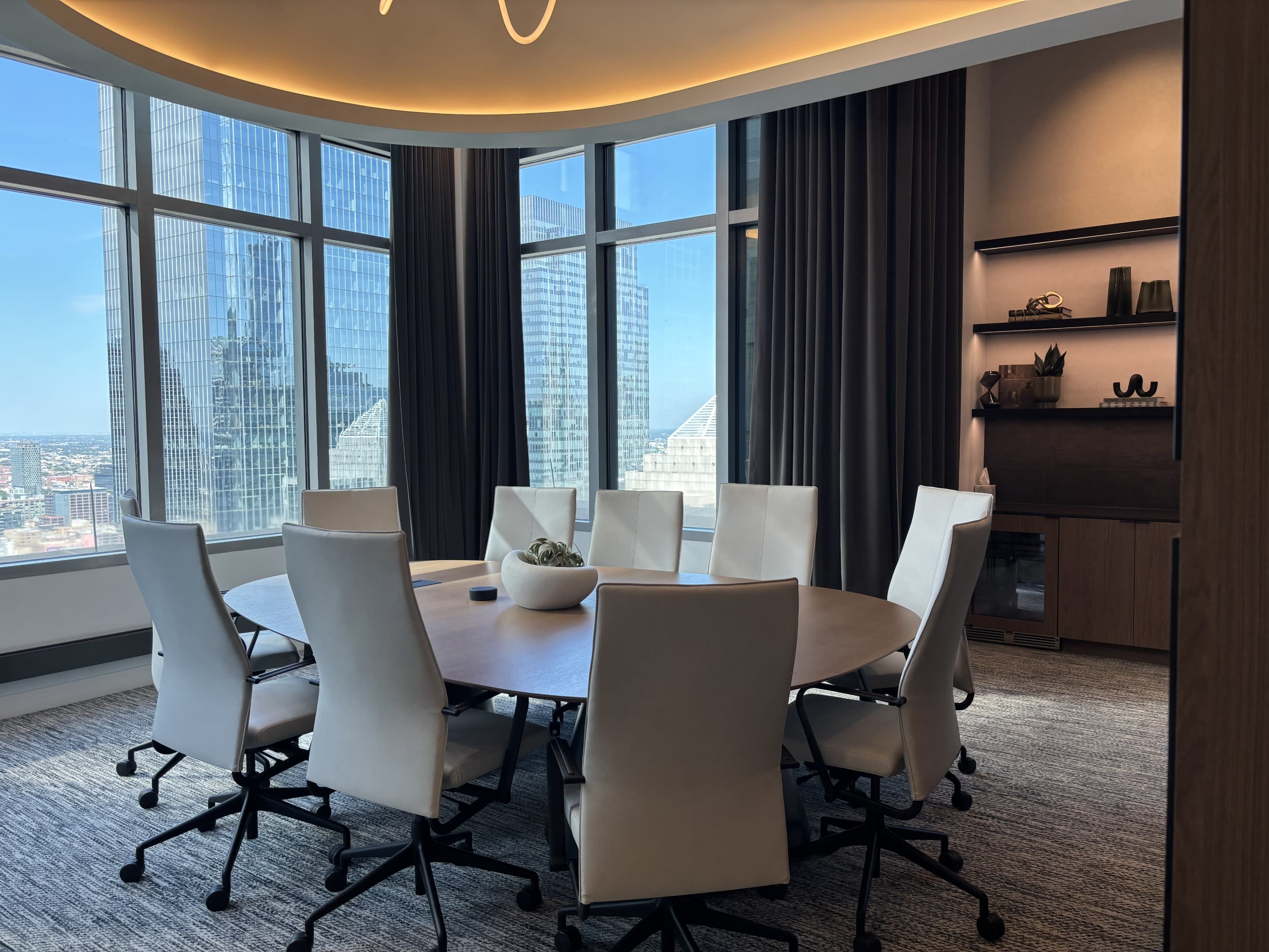 The image shows a modern conference room with a round table surrounded by white chairs, large windows offering a view of the city skyline, and contemporary decor.