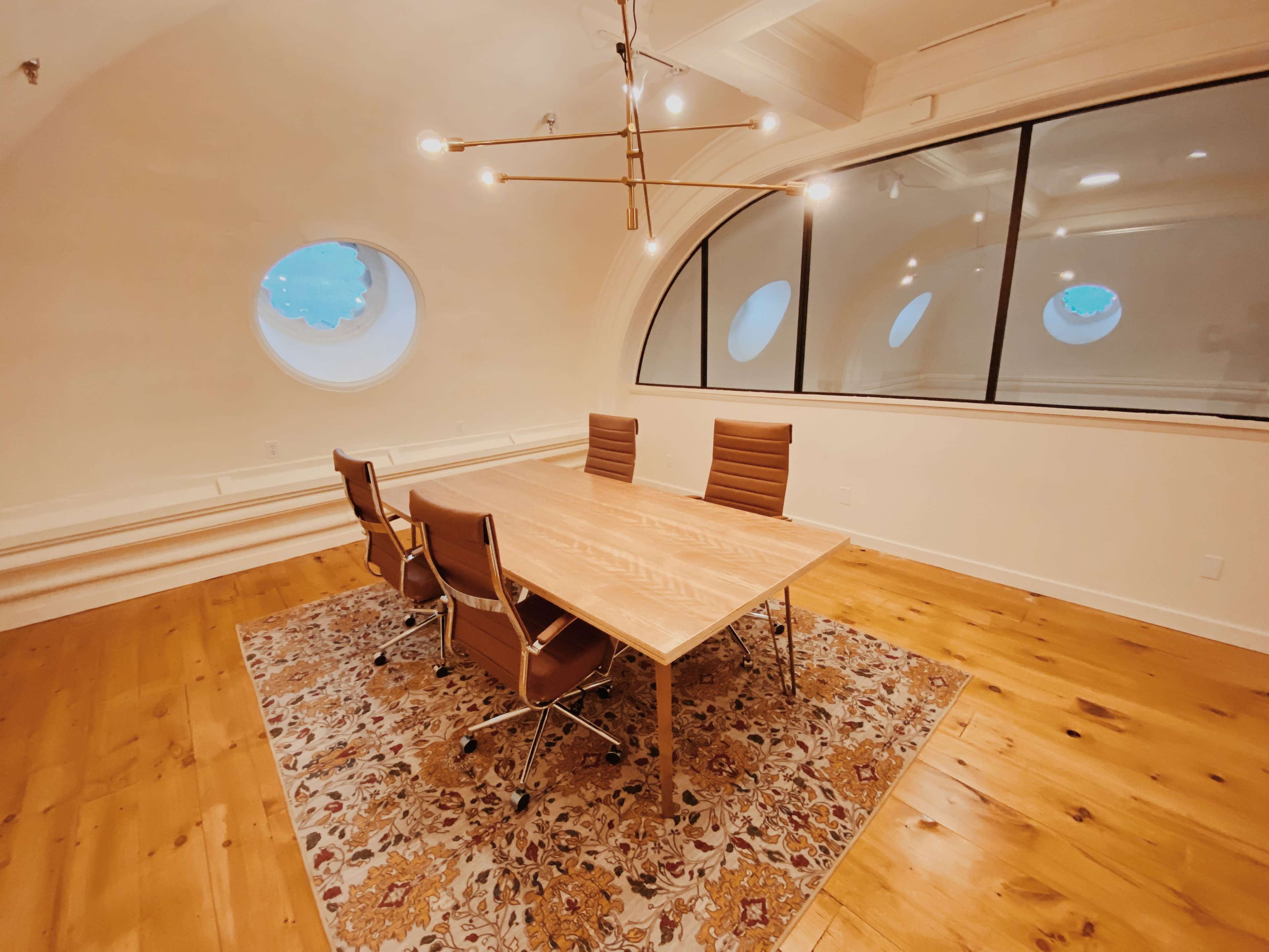 A modern conference room features a wooden table surrounded by four brown leather chairs, with large circular windows and wooden flooring.