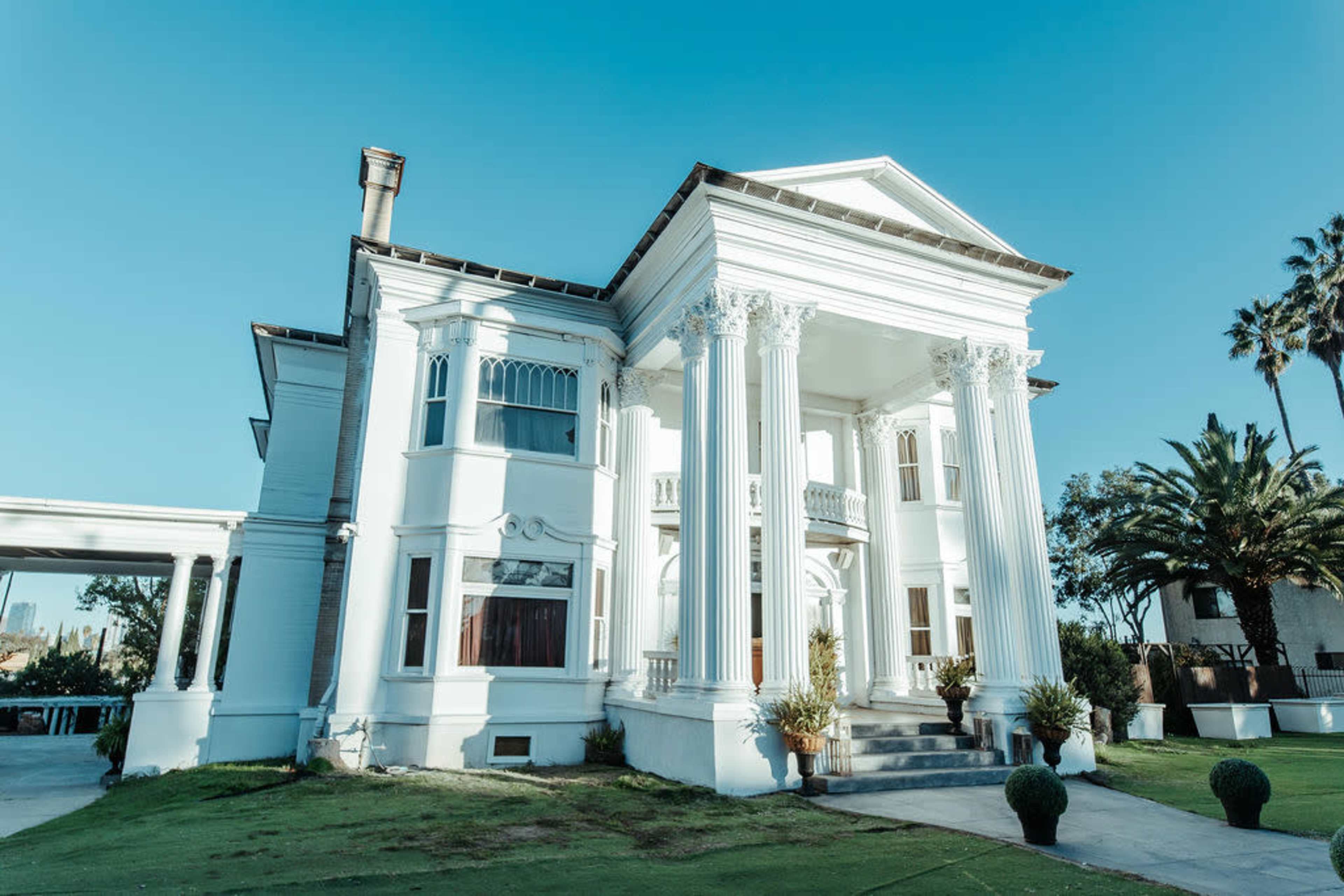 The image shows a large, white mansion with decorative columns and a well-maintained lawn, surrounded by palm trees under a clear blue sky.