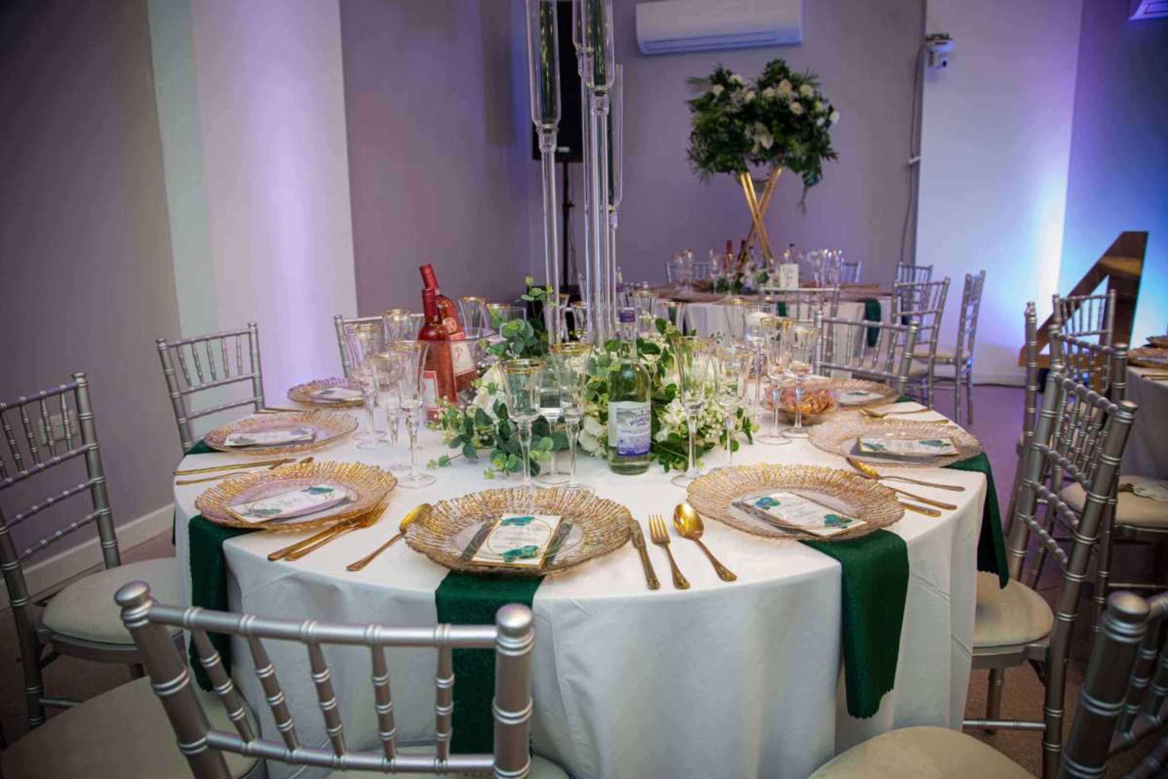 The image shows a elegantly set dining table adorned with gold plates, glassware, and green accents, surrounded by silver chiavari chairs.