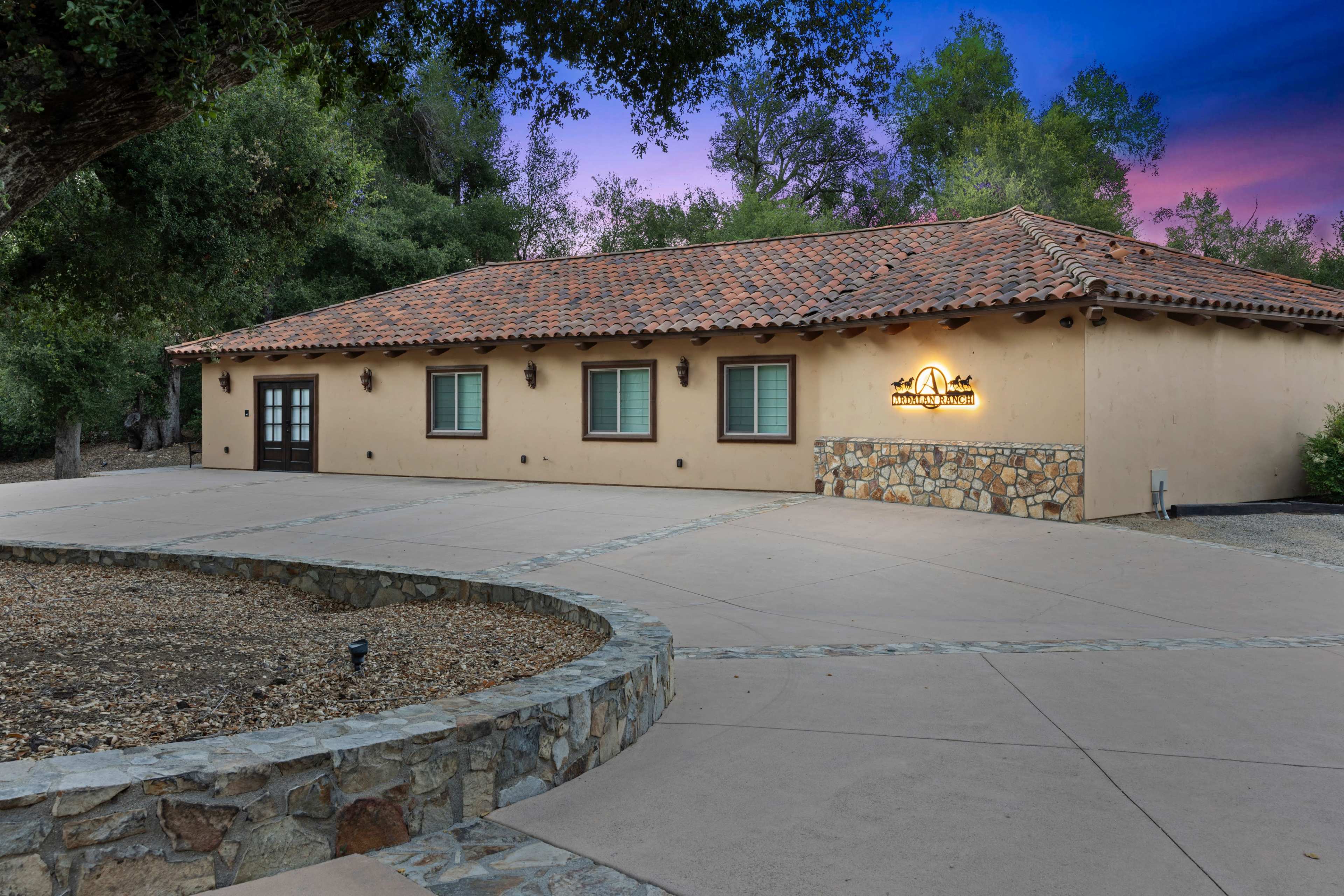 A single-story adobe-style building with a tiled roof and stone accents sits in a landscaped area under a colorful sky.
