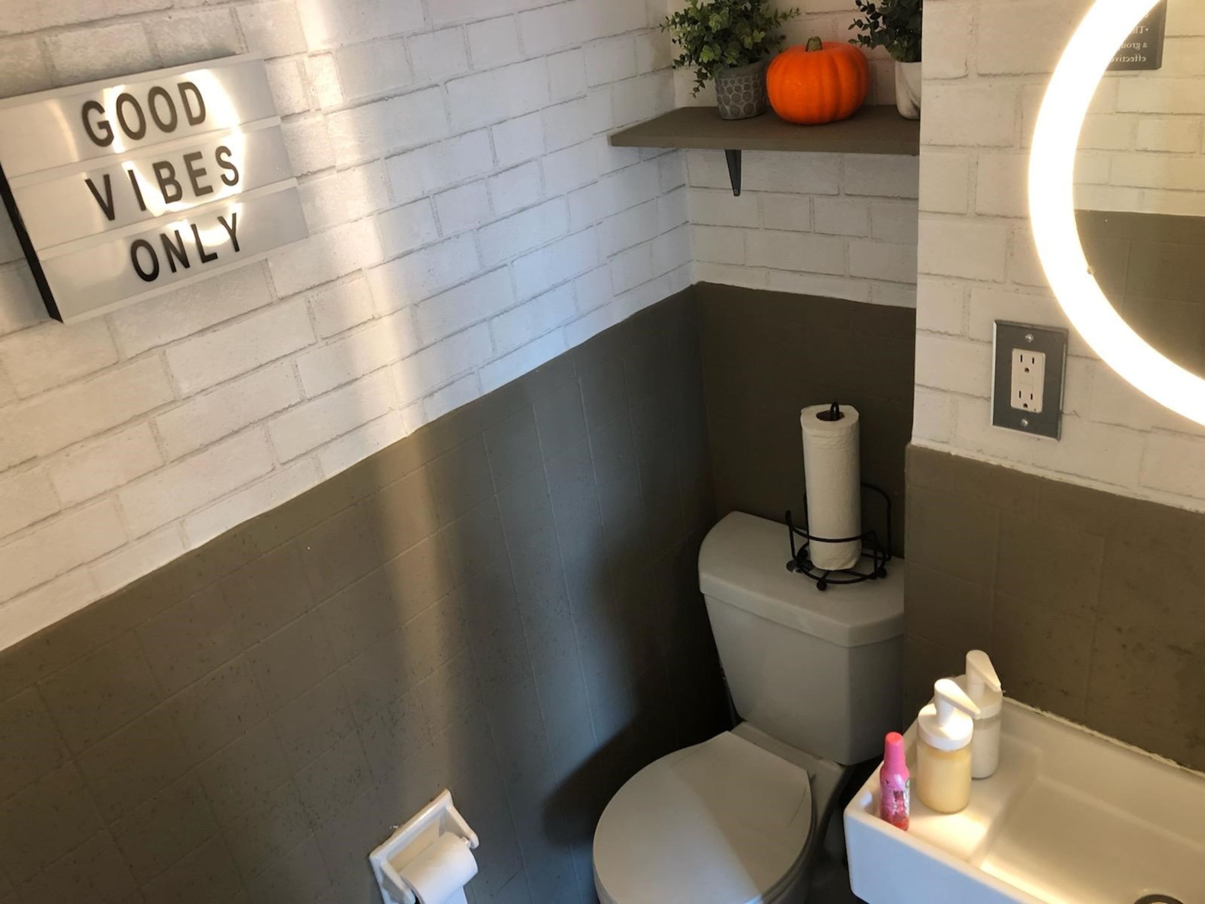 A small bathroom features a white brick wall, a decorative shelf with plants and a pumpkin, and a lighted sign that reads "GOOD VIBES ONLY."