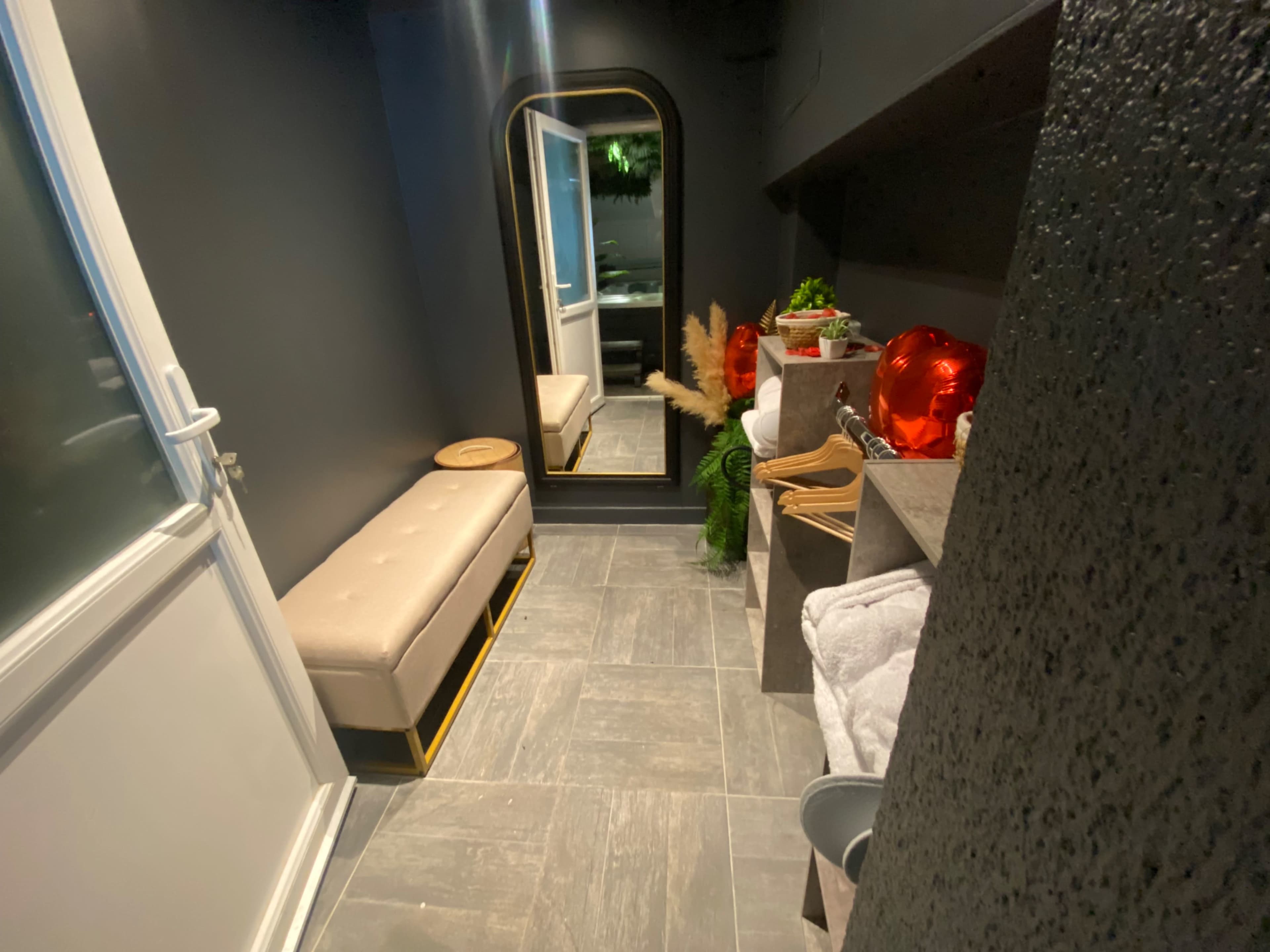 A narrow hallway with a bench, a large mirror, and shelves containing plants and towels against dark walls.
