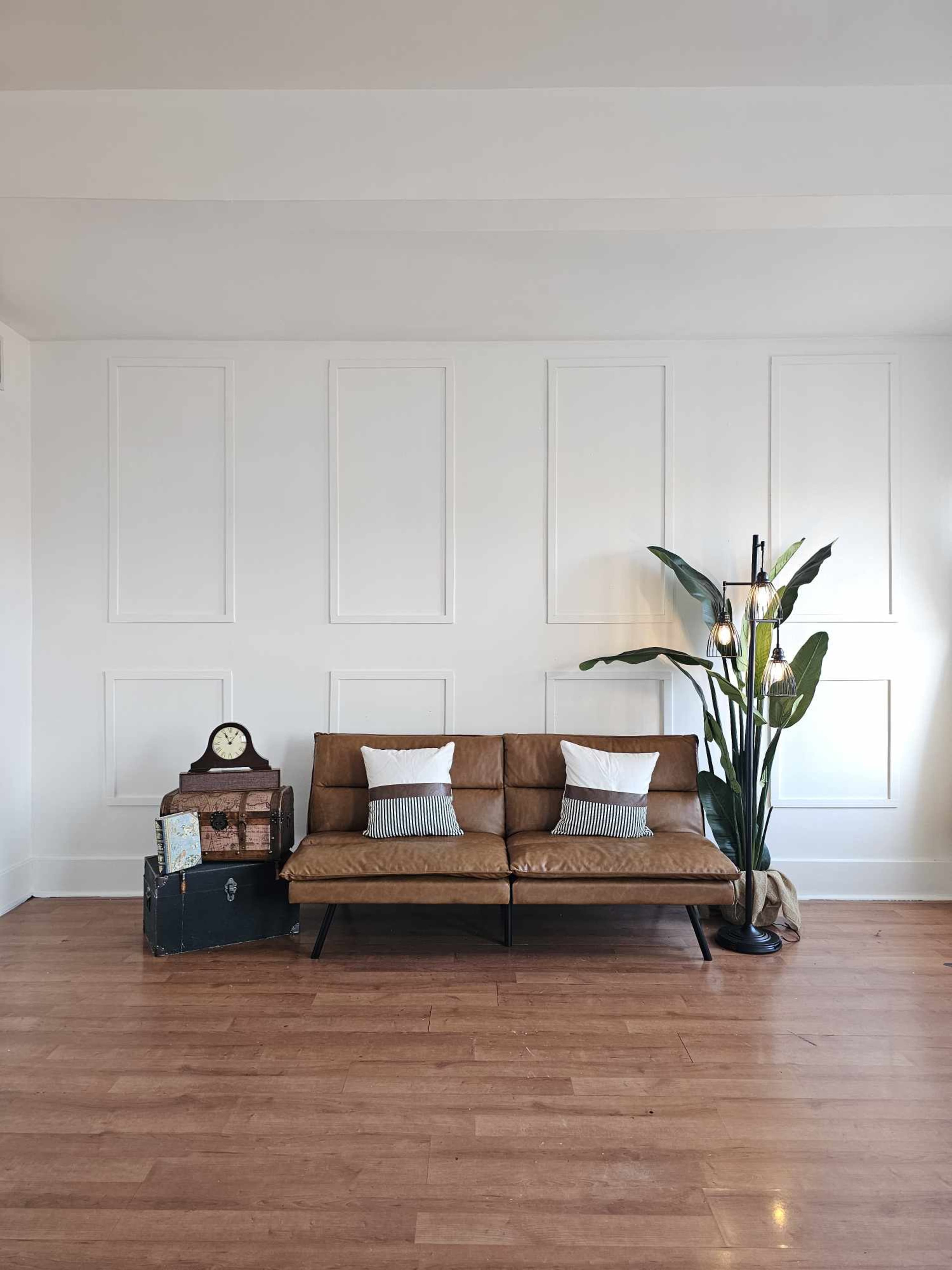A mid-century leather sofa with white and striped pillows is positioned in a bright room with a large plant, a vintage clock, and wooden flooring.