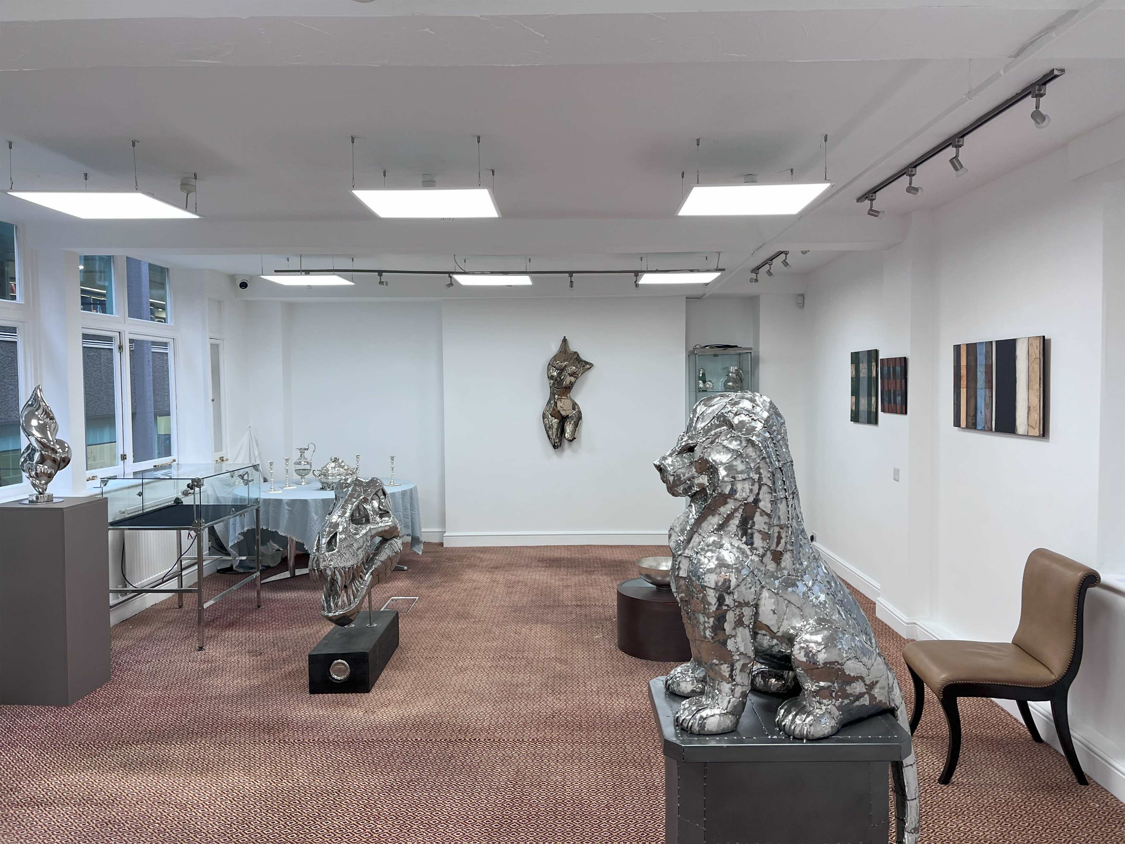 The image shows a modern art gallery featuring metallic sculptures of a lion and a figure, along with several abstract paintings on the walls and a glass display case.