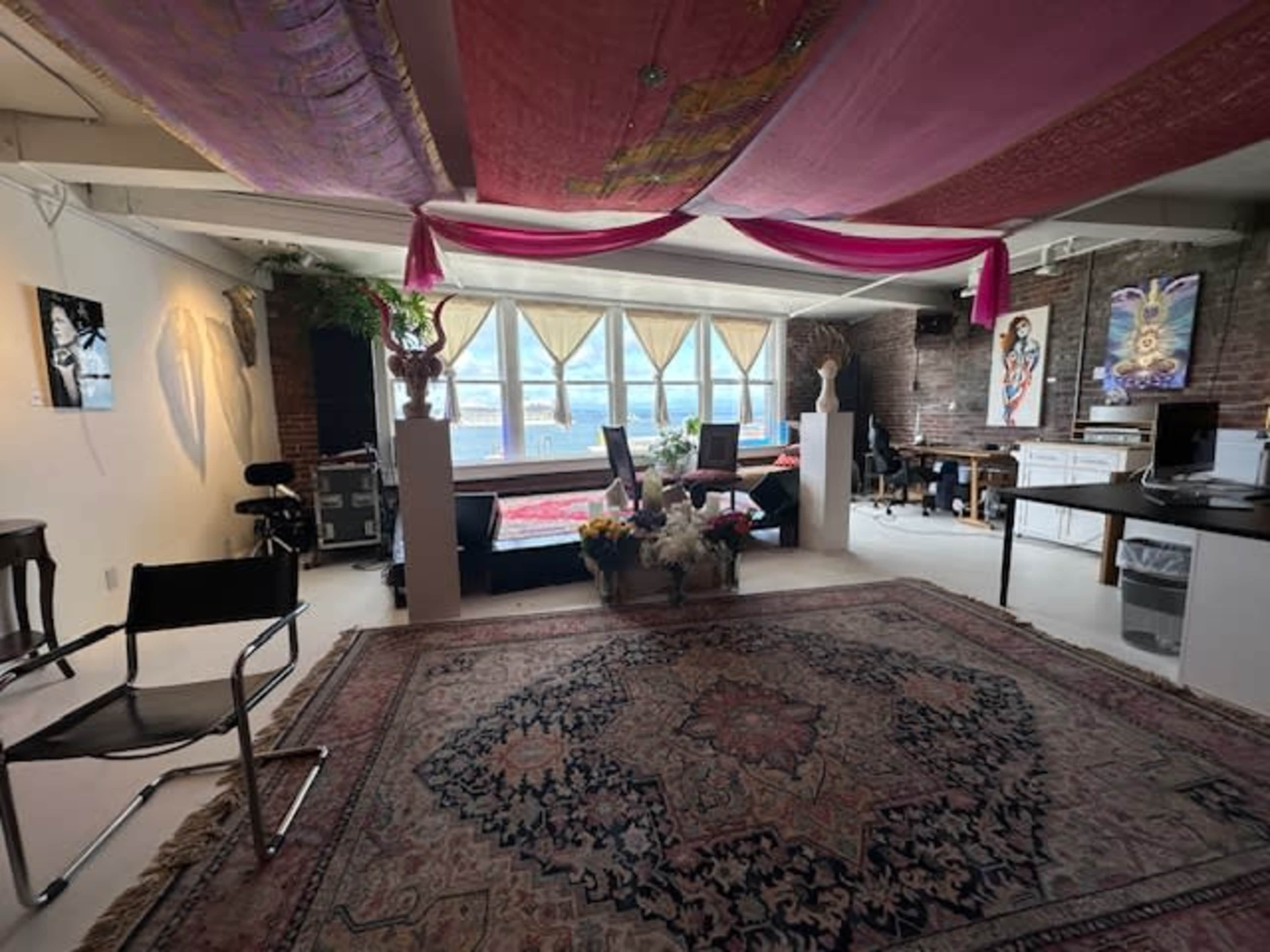 A spacious room with large windows, featuring a decorative rug, two chairs, a table, and art on the walls.