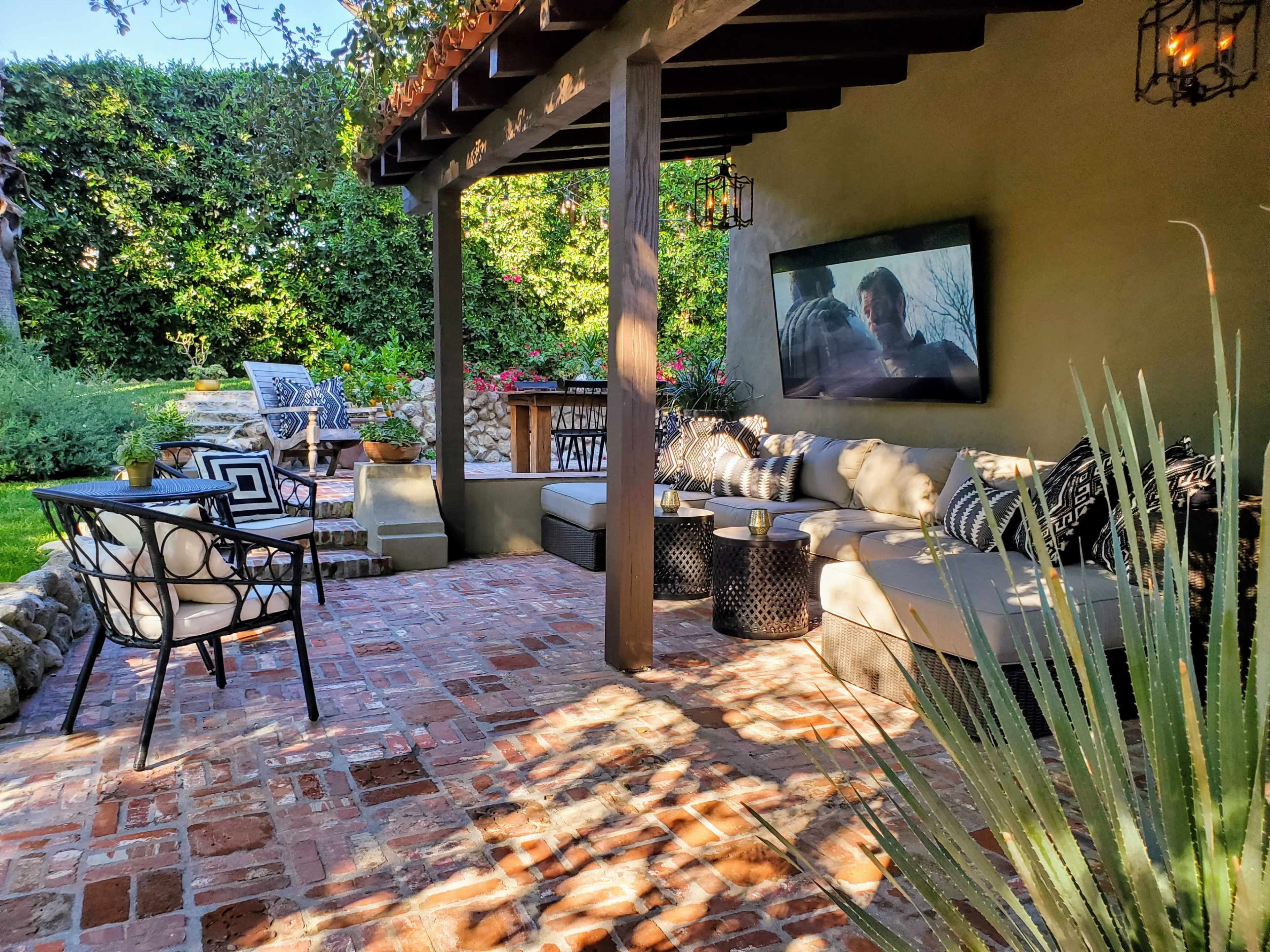 A covered patio with a sectional couch, a large television mounted on the wall, and various seating arrangements surrounded by greenery.