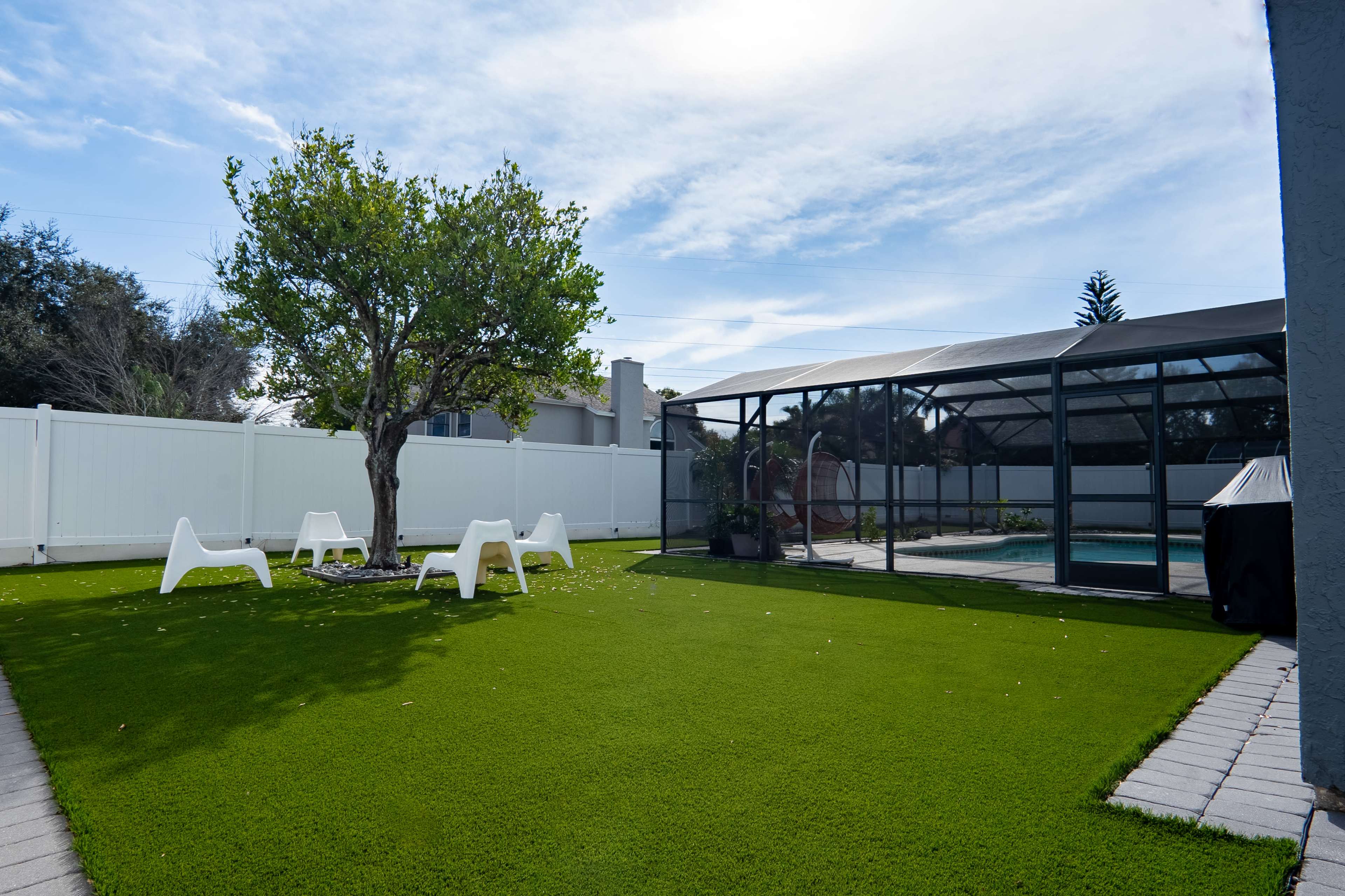 A well-maintained backyard features artificial grass, a tree, white chairs, and a screened pool area.