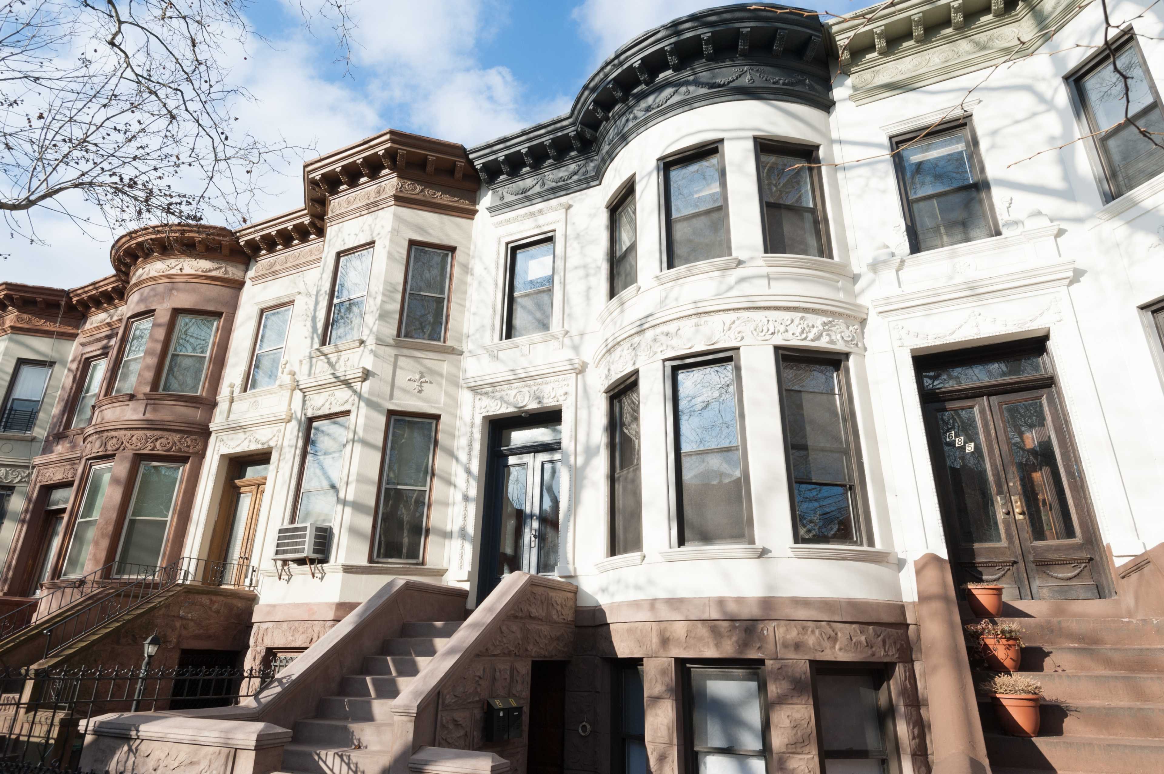 A row of brownstone buildings features a mix of colored facades and decorative architectural details under a partly cloudy sky.