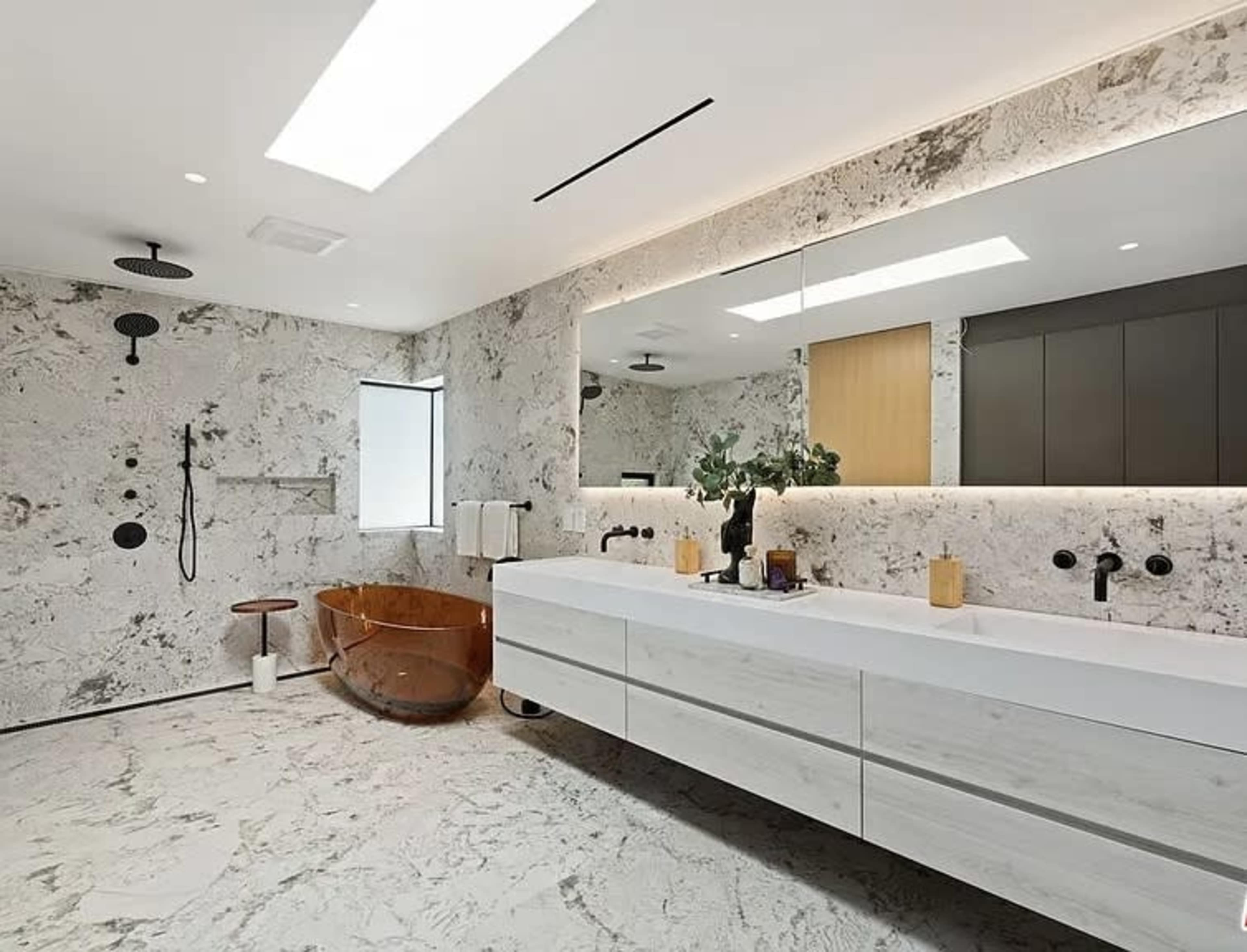 A modern bathroom features a freestanding copper bathtub, a double vanity with a white countertop, and large marble-patterned tiles on the walls and floor.