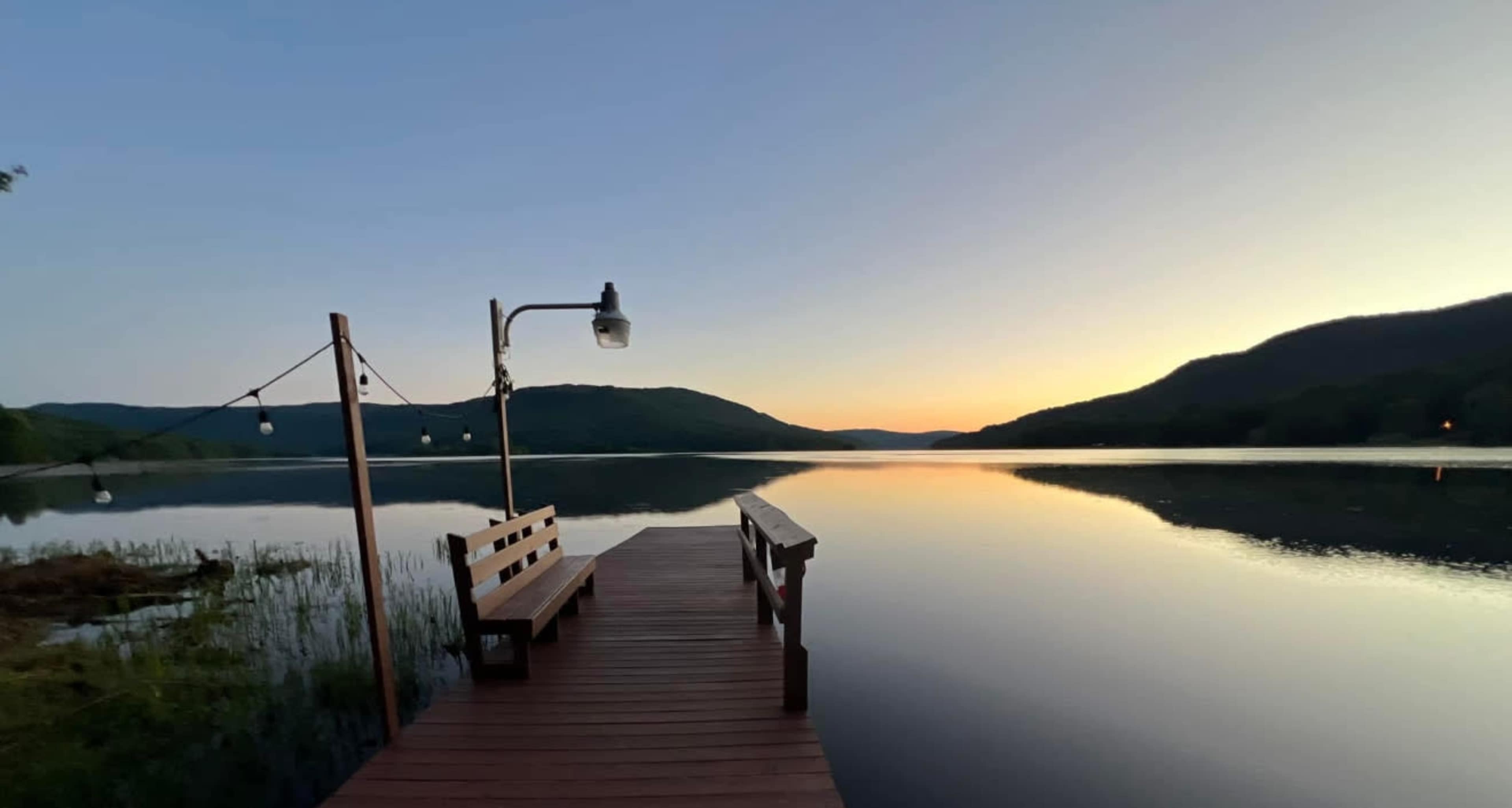 A wooden dock extends into a calm lake, surrounded by mountains, with string lights hanging overhead against a backdrop of a colorful sunset.