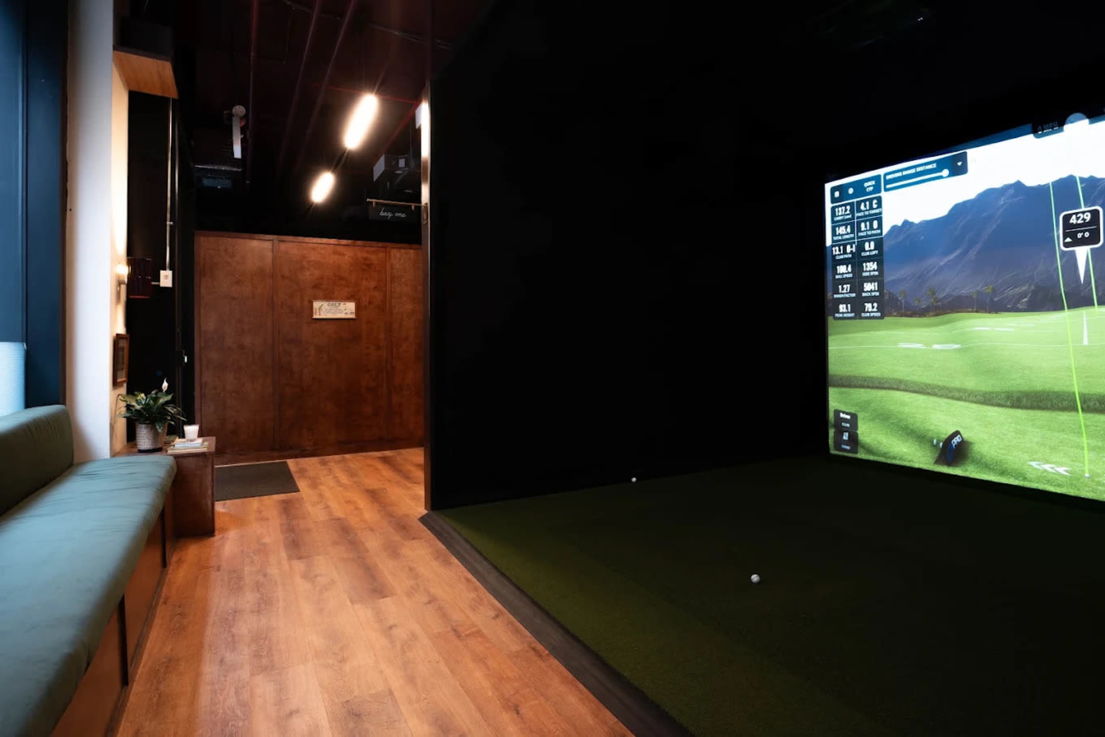 The image shows a modern indoor golfing simulator room featuring a putting green and a large screen displaying a golf course.