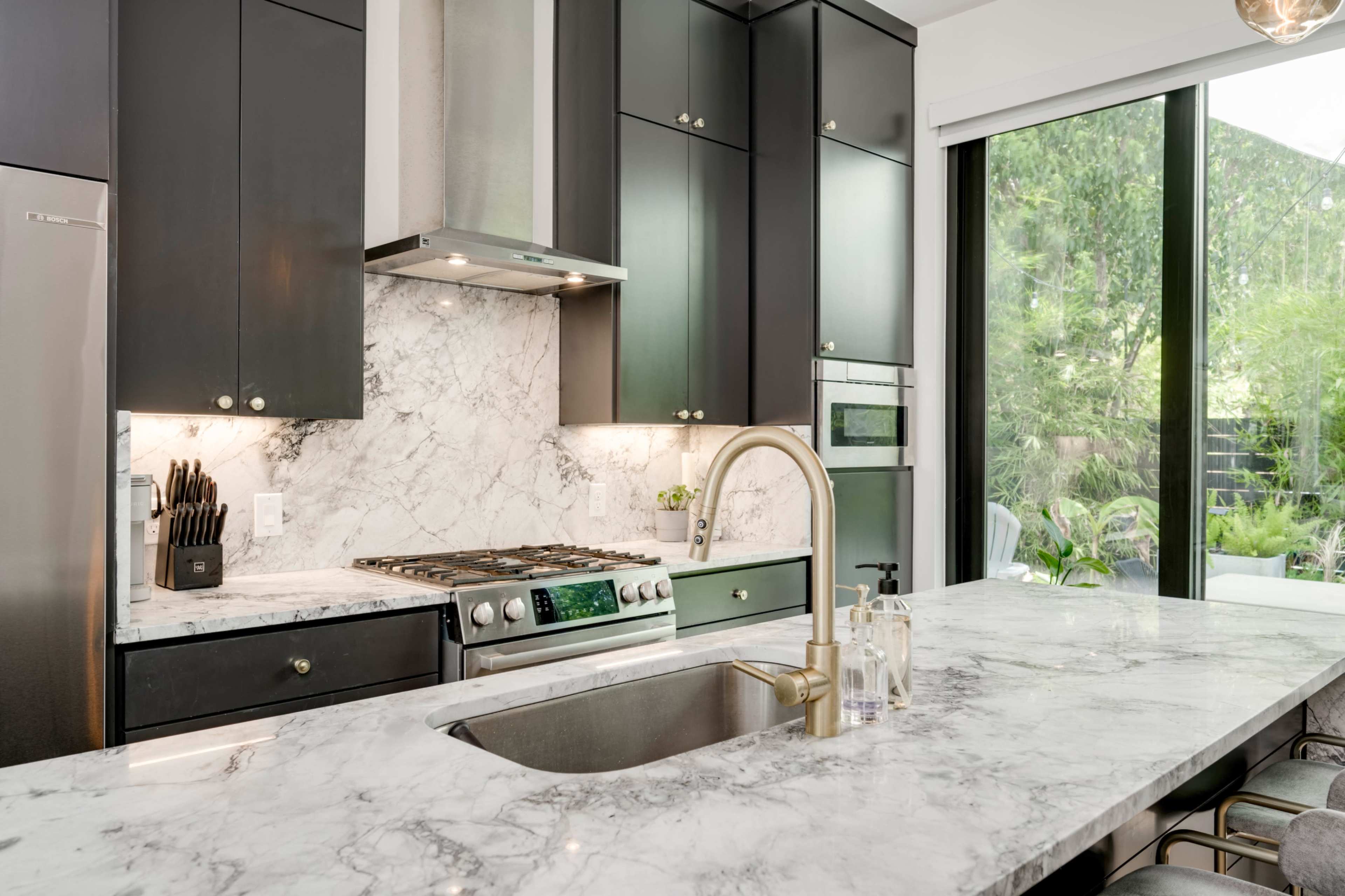 The image features a modern kitchen with black cabinets, a marble countertop, and a large window showcasing greenery outside.