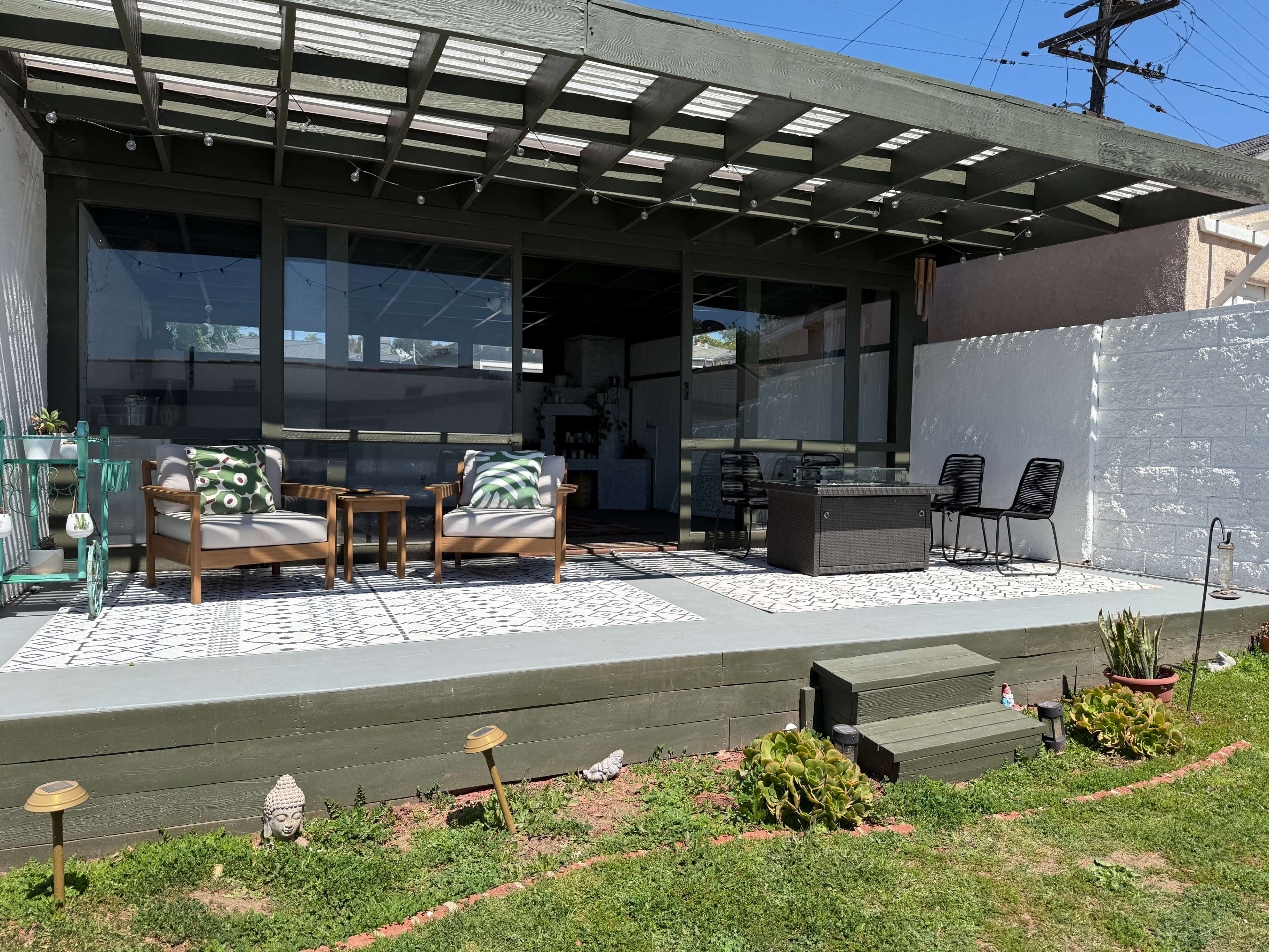 The image shows a covered patio area with seating, a small table, and a decorative lawn, surrounded by white walls and greenery.