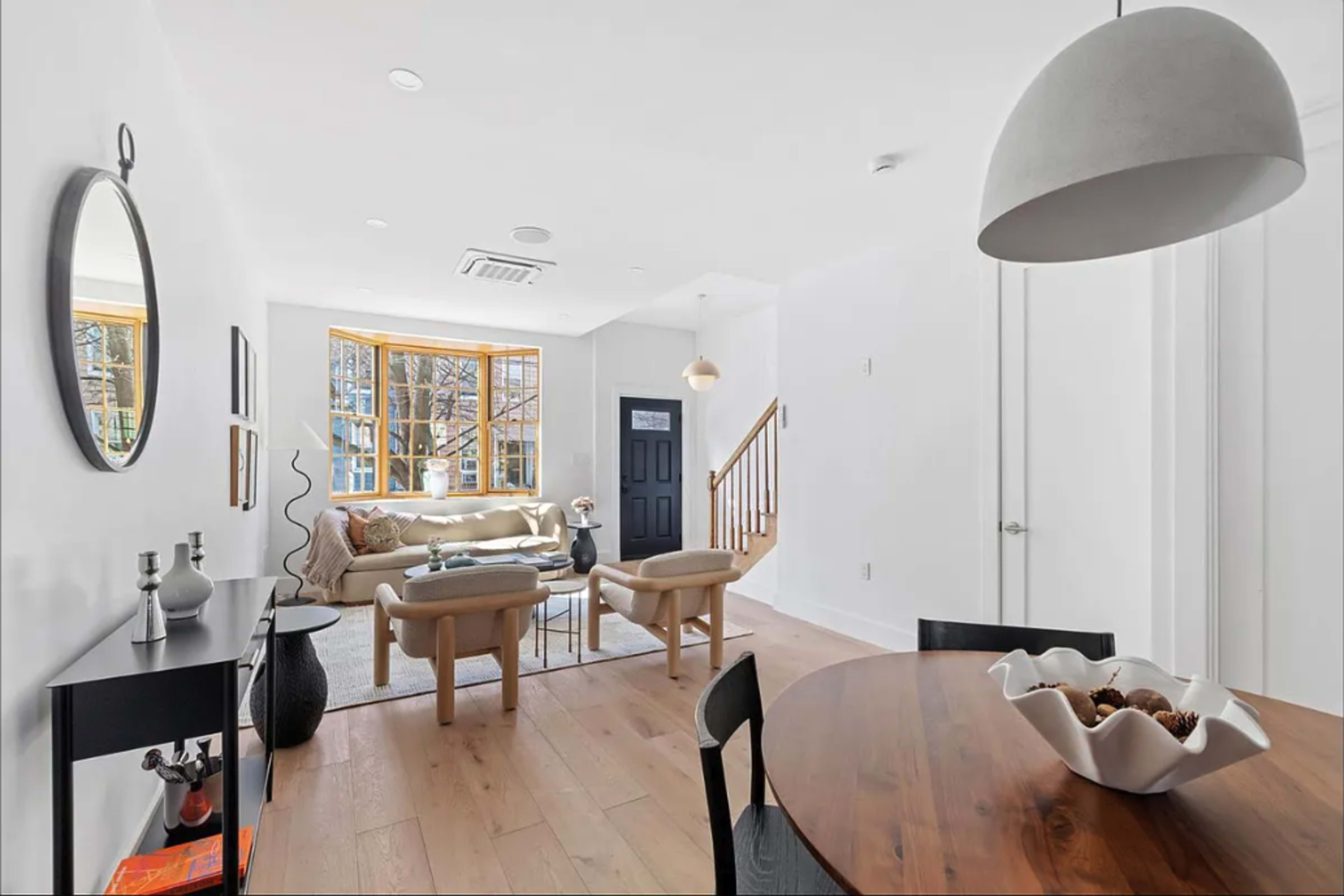 The image shows a modern living space with a beige sofa, a wooden dining table, and large windows that let in natural light.