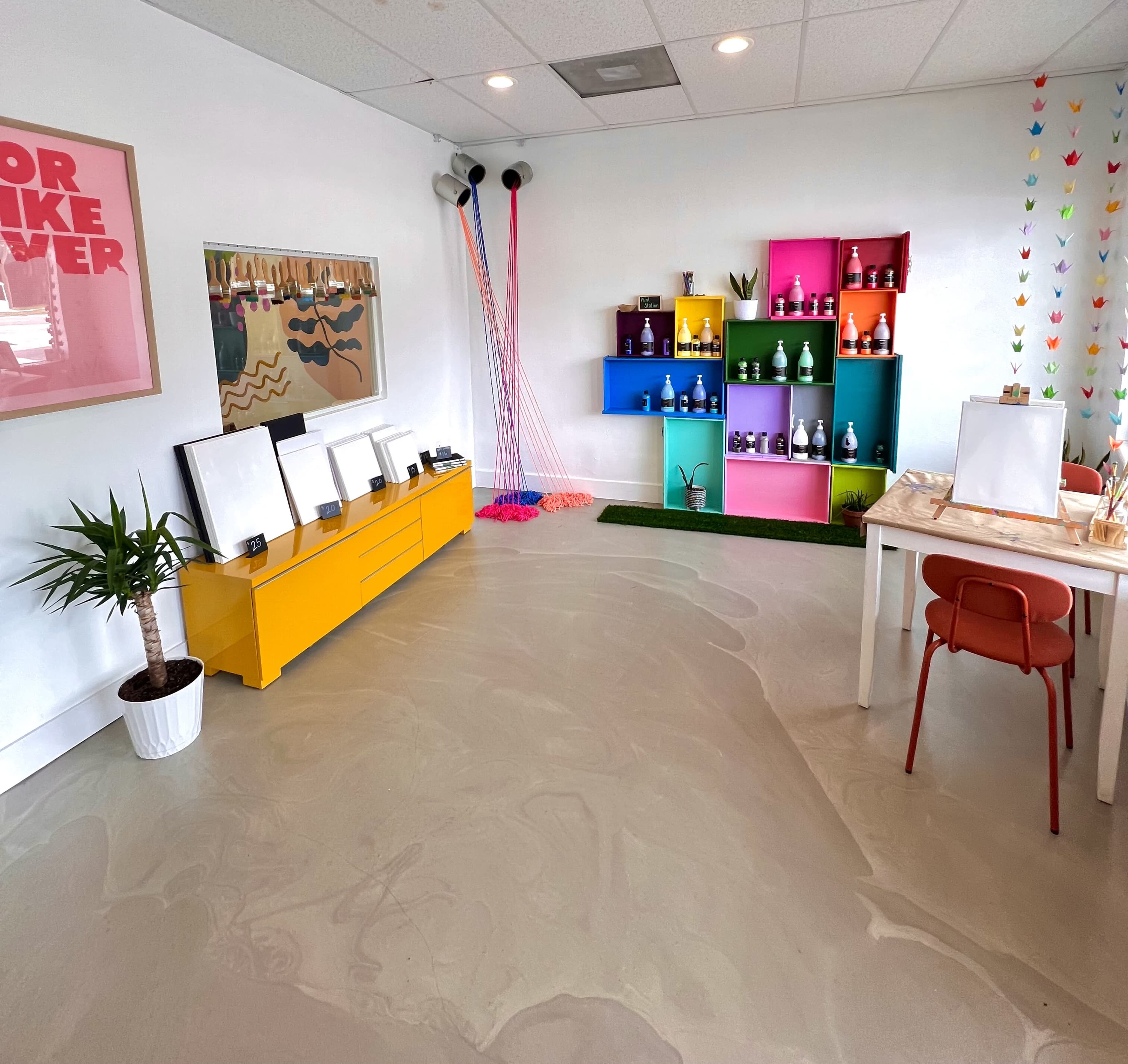 A brightly colored room with a yellow cabinet, a white table and chairs, plants, and shelves displaying various bottles against a light-colored floor.