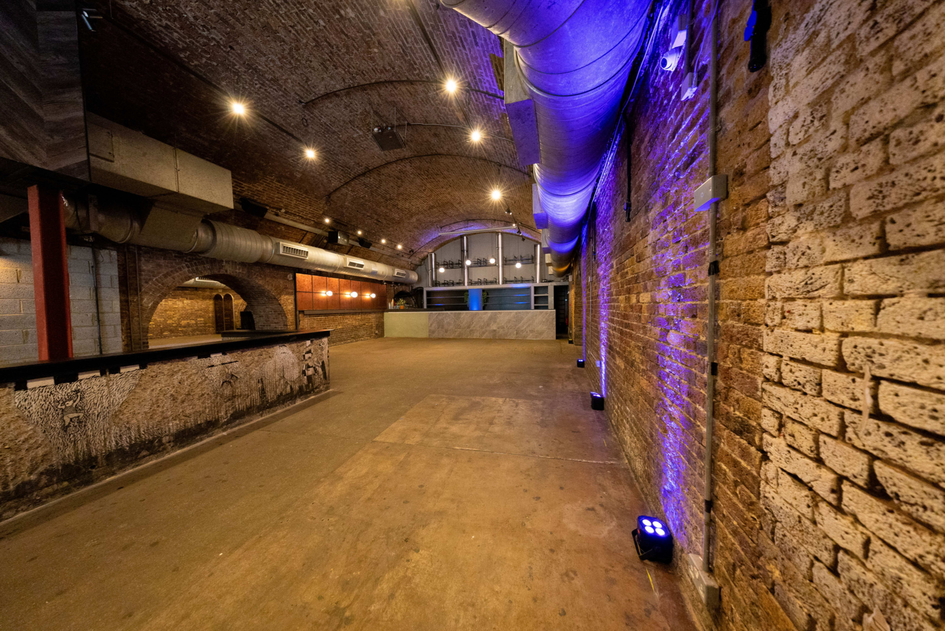 The image shows an empty industrial-inspired venue with exposed brick walls, a bar area, and soft lighting.