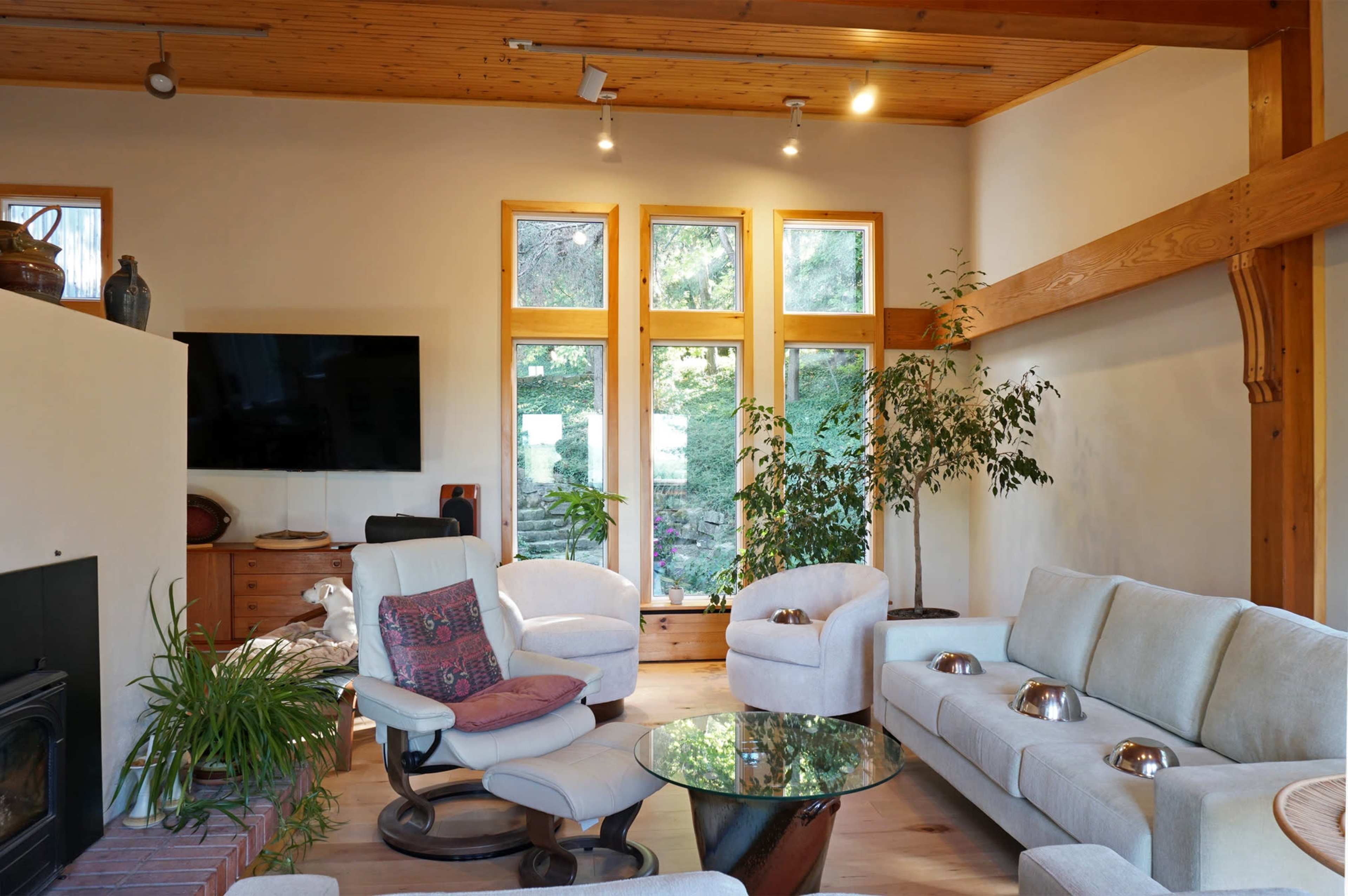 The image shows a modern interior living room featuring large windows, a circular glass coffee table, and white furniture arranged around a fireplace.