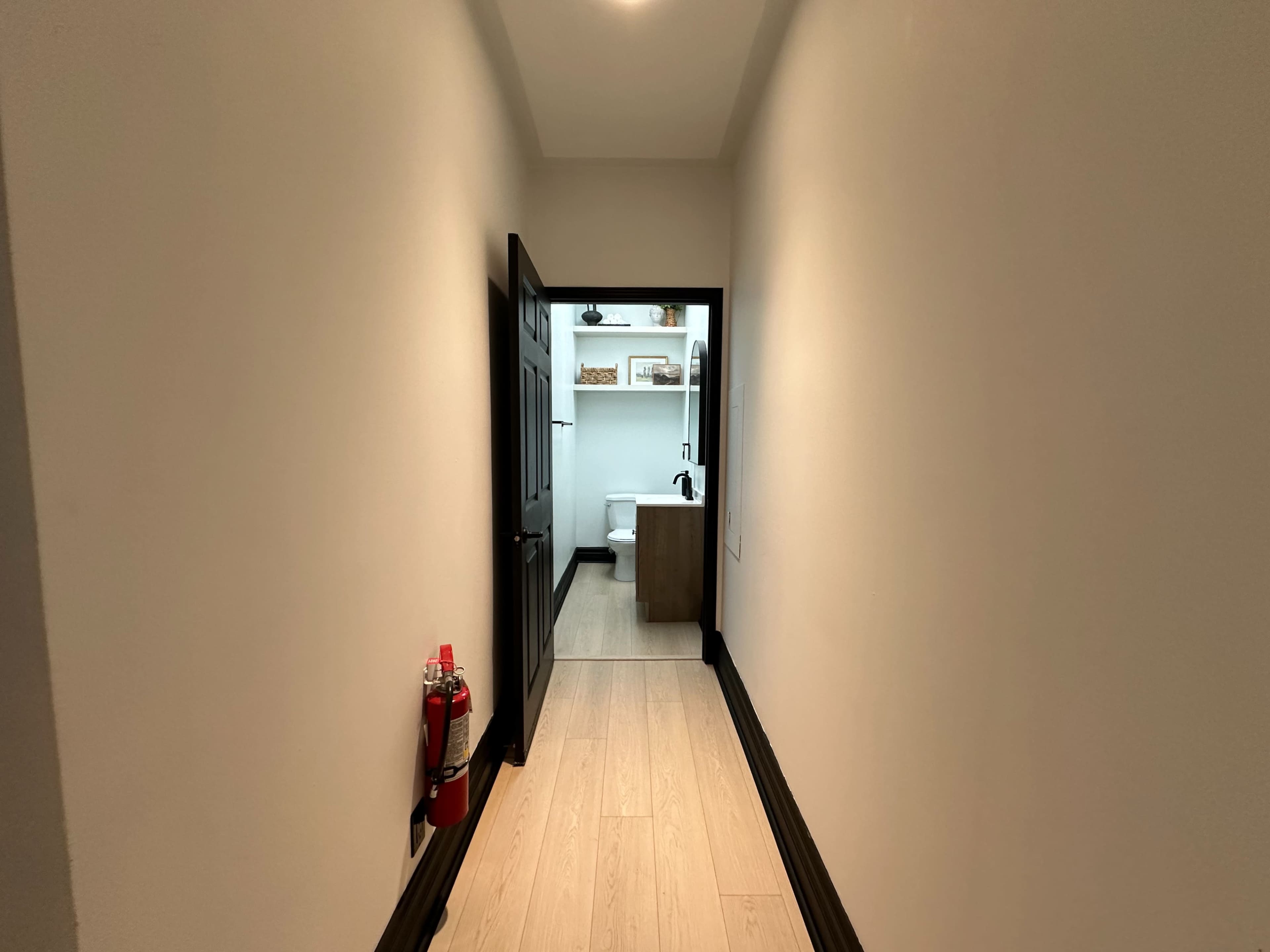 A narrow hallway with light-colored wooden floors, leading to a bathroom visible at the end.