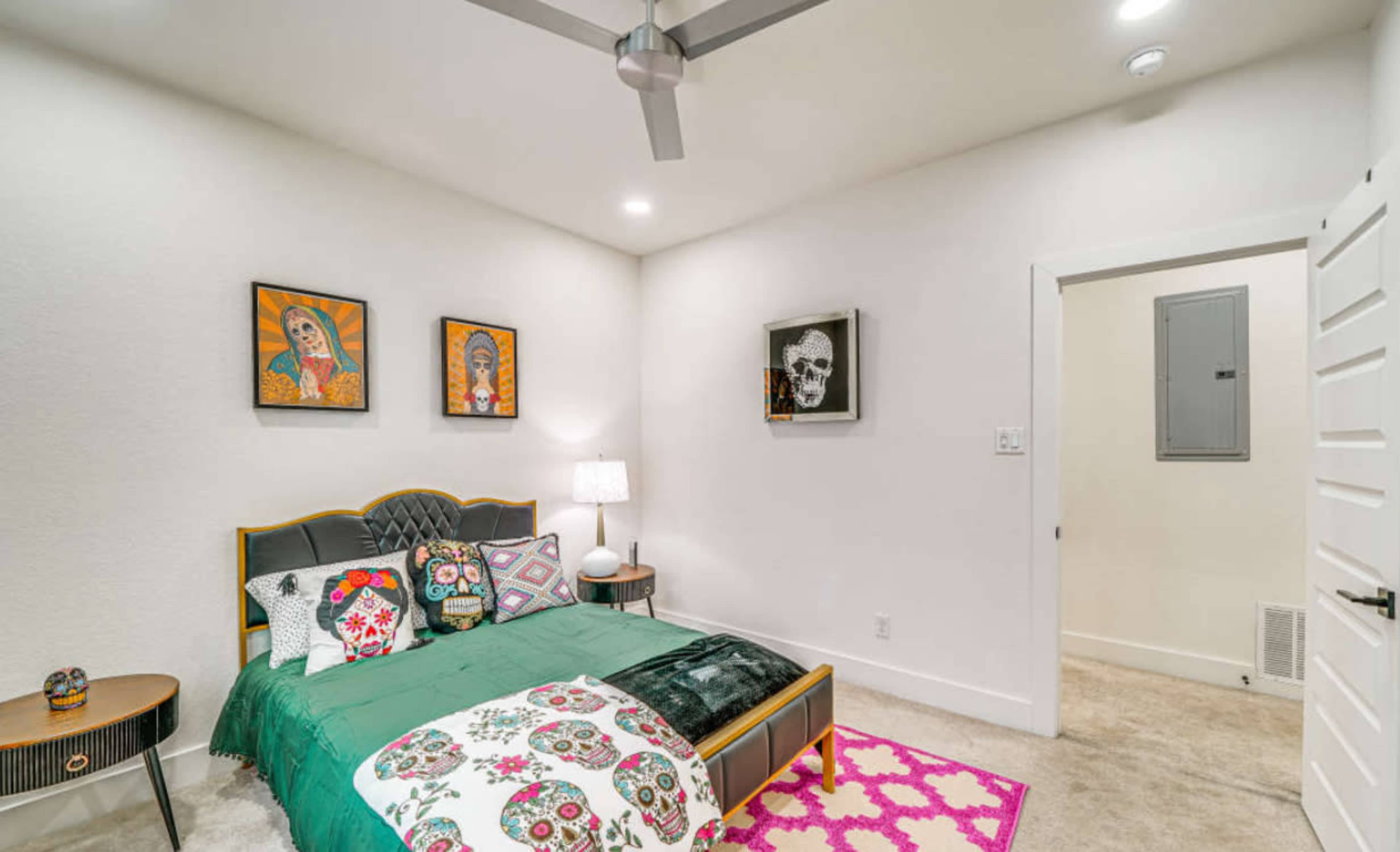 The image shows a decorated bedroom featuring a bed with colorful bedding, wall art of skulls and figures, a nightstand with a lamp, and a light-colored rug.