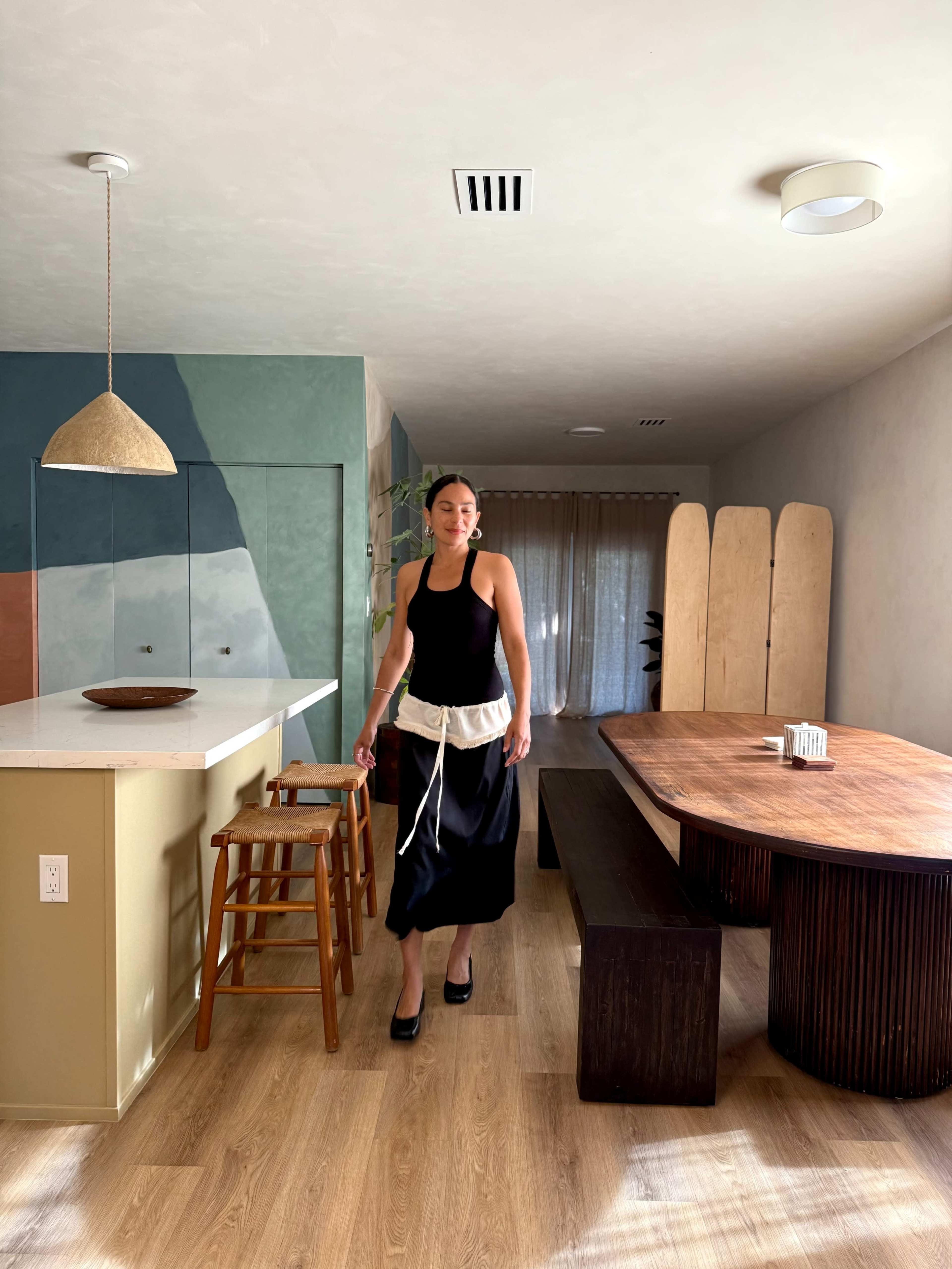 A woman in a black dress and white apron walks through a modern, minimally decorated kitchen and dining area with wooden furniture and colorful wall accents.