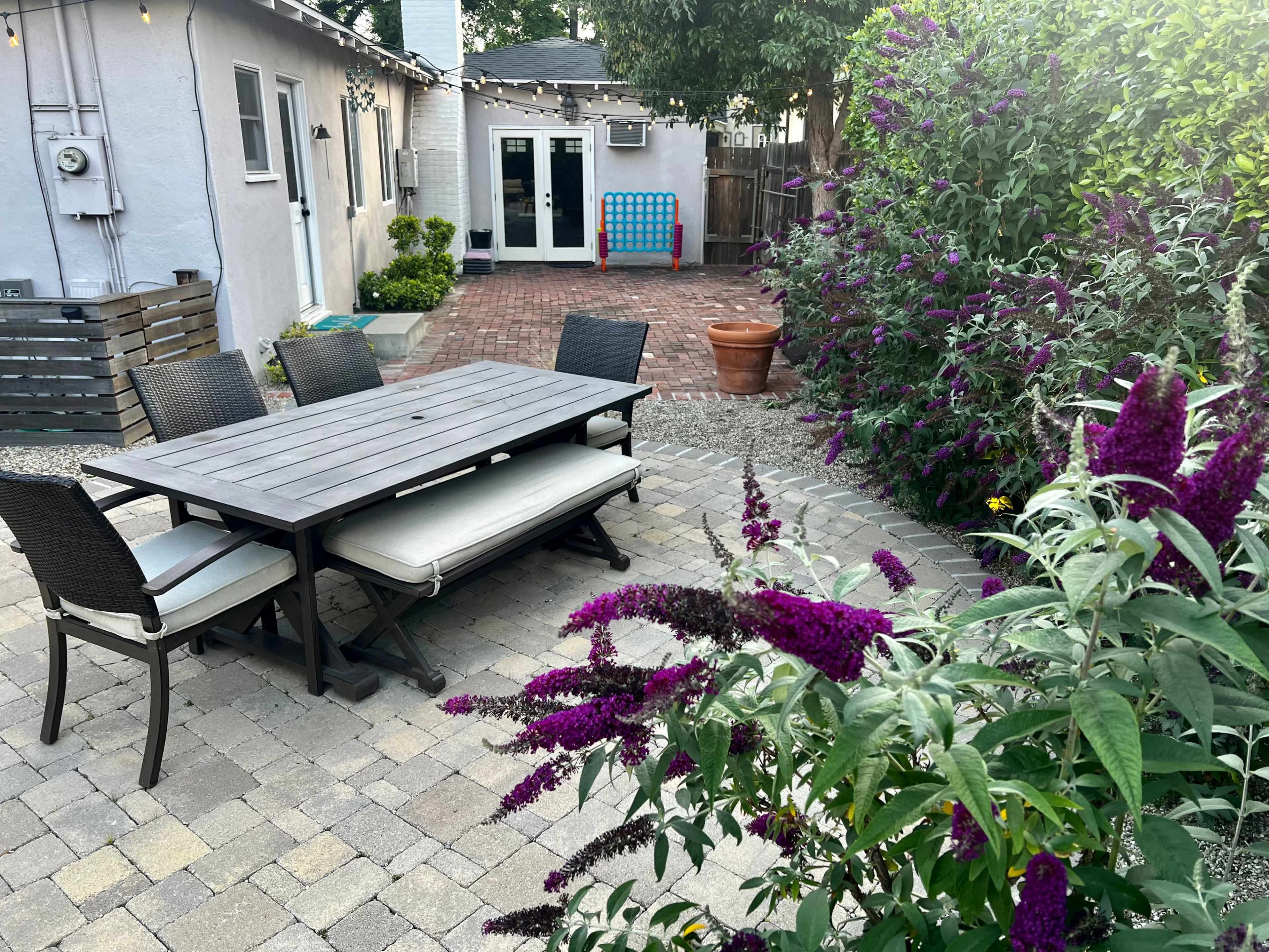 A wooden dining table with chairs is positioned on a stone patio surrounded by flowering plants and a brick pathway leading to a nearby building.