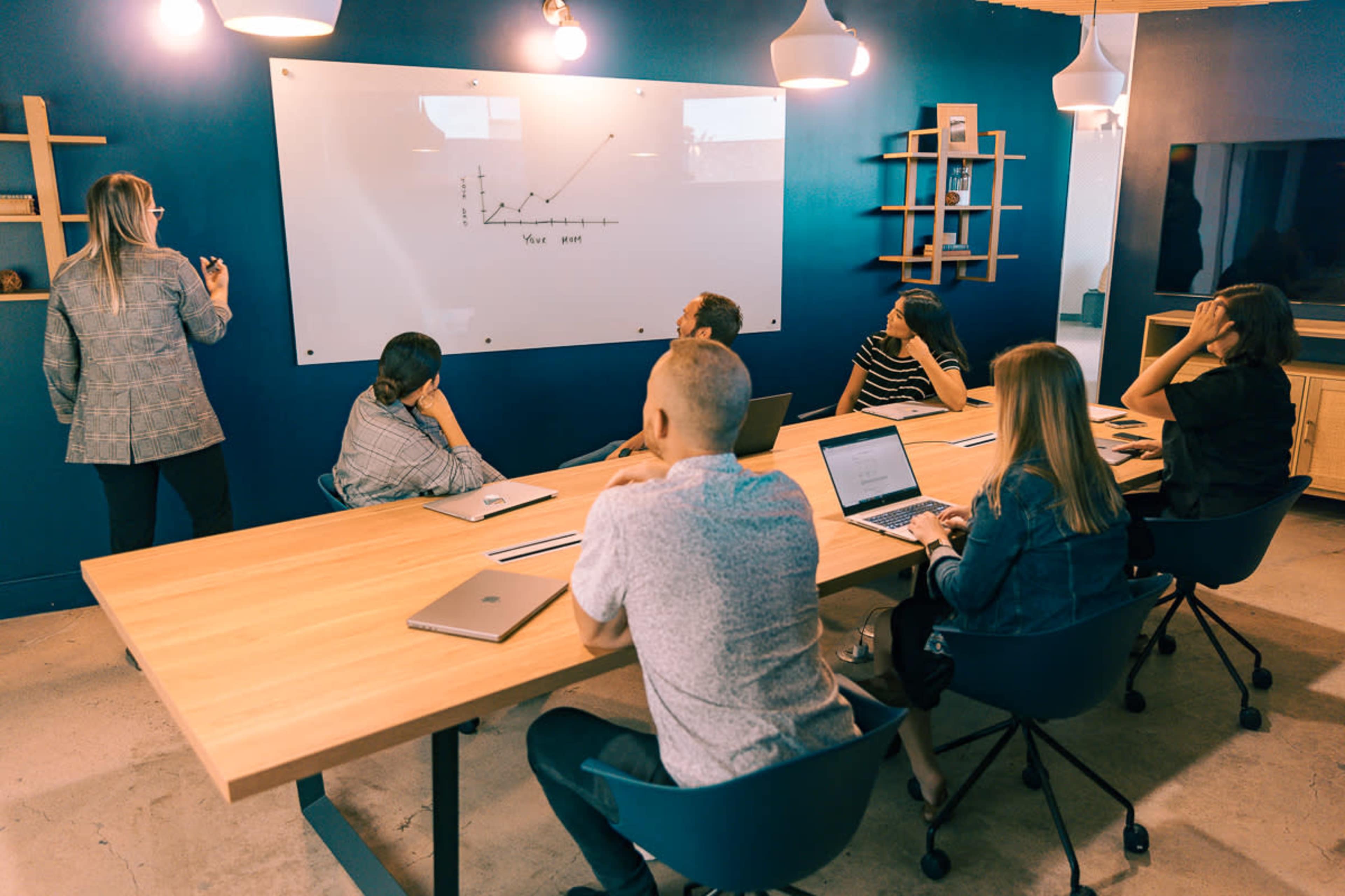 A group of professionals sits around a conference table while one person presents information on a whiteboard.