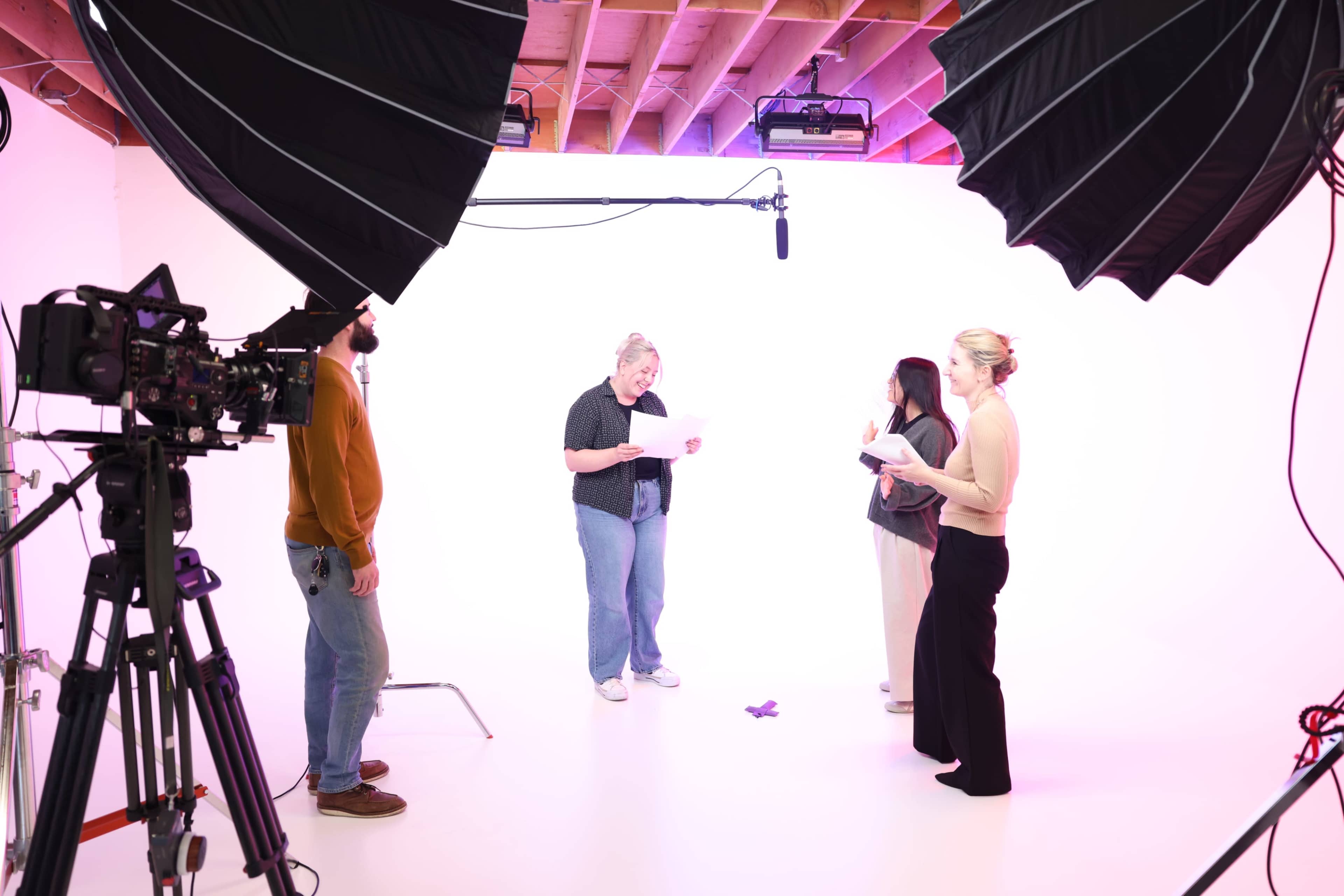 A group of four people is gathered in a well-lit studio, reading from scripts while two large softboxes and a microphone on a boom are positioned overhead.