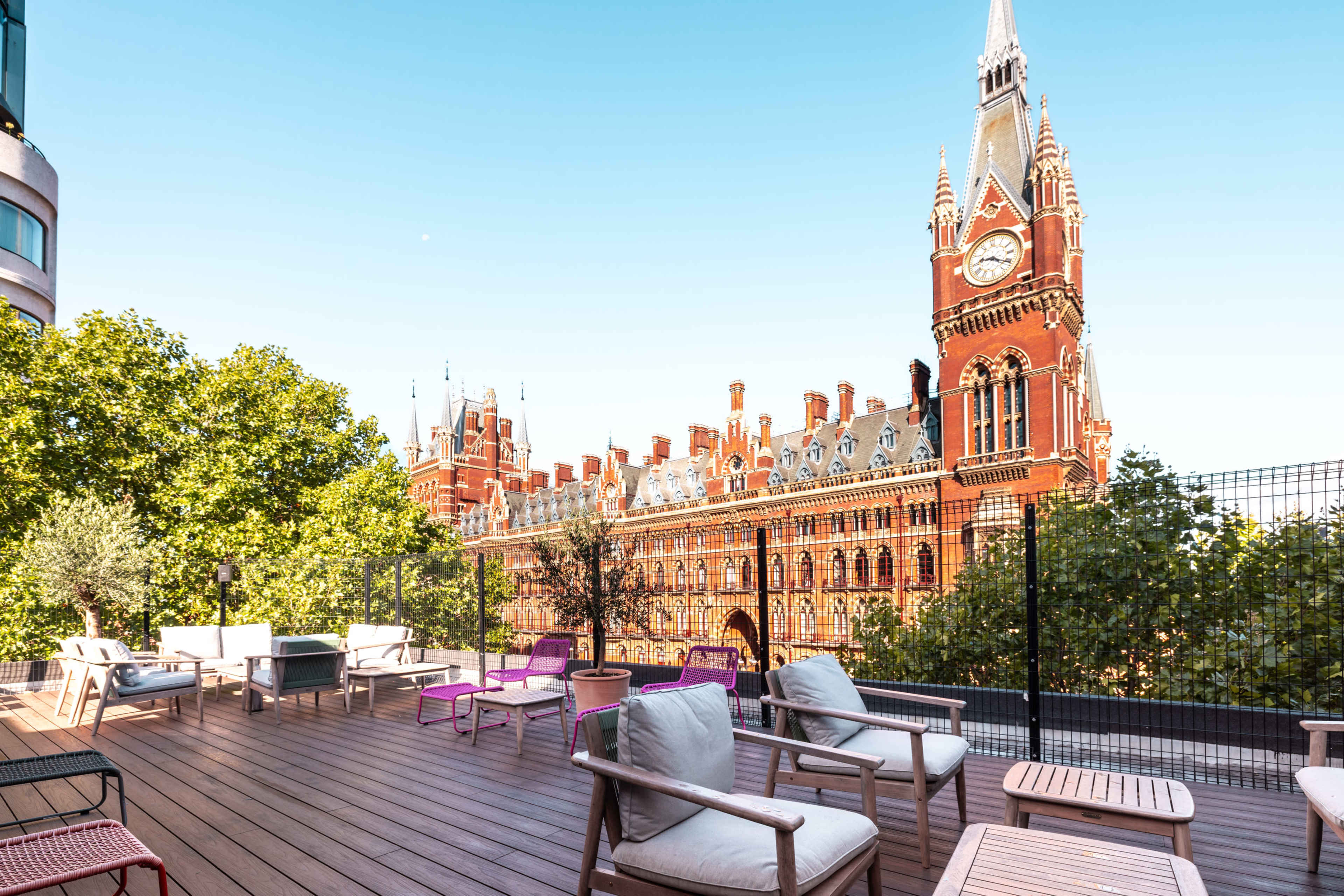 The image shows a rooftop terrace with wooden flooring and seating that overlooks a historic red brick building with a clock tower.