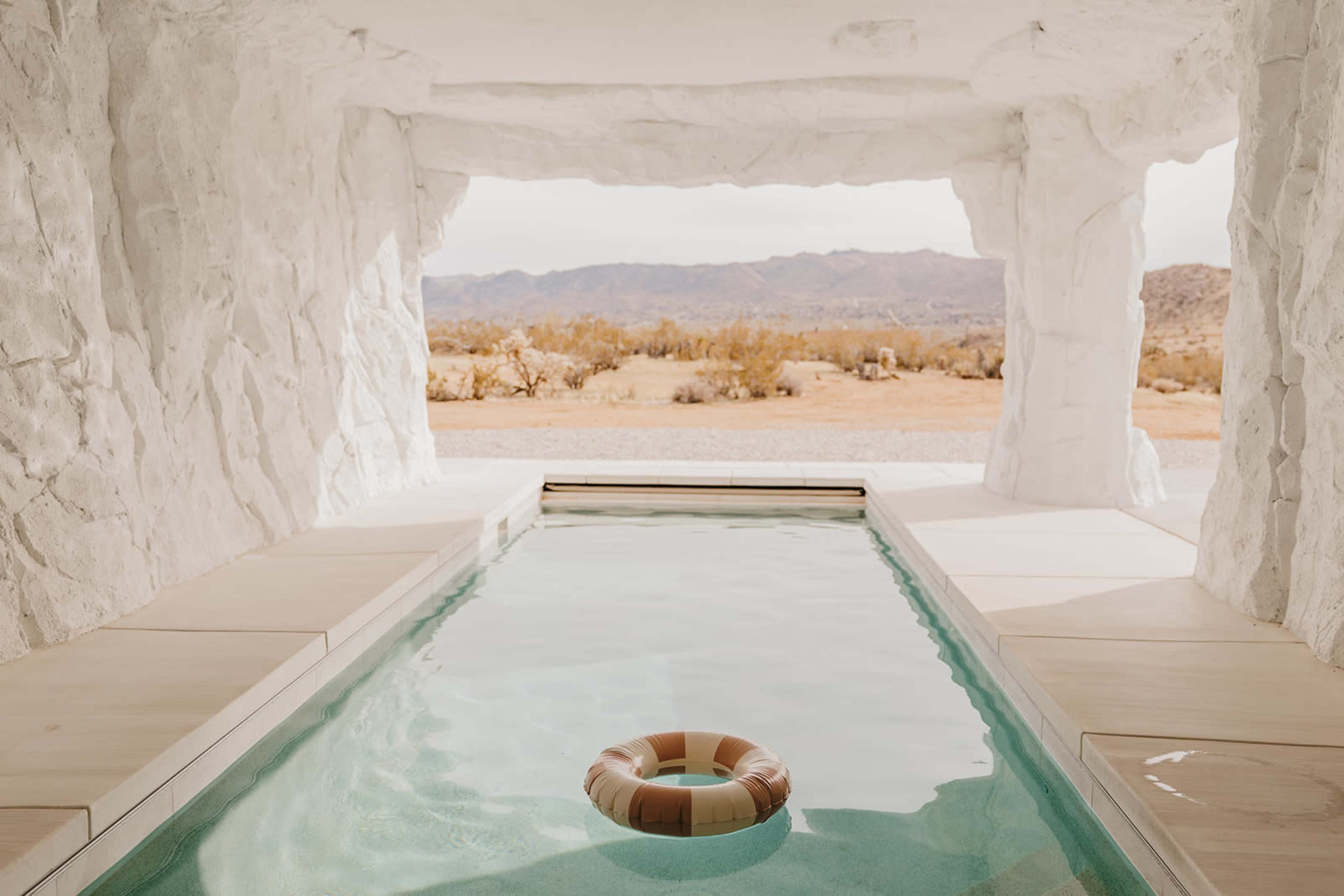 A stone-walled room features a swimming pool with a single inflatable ring, looking out onto a desert landscape.