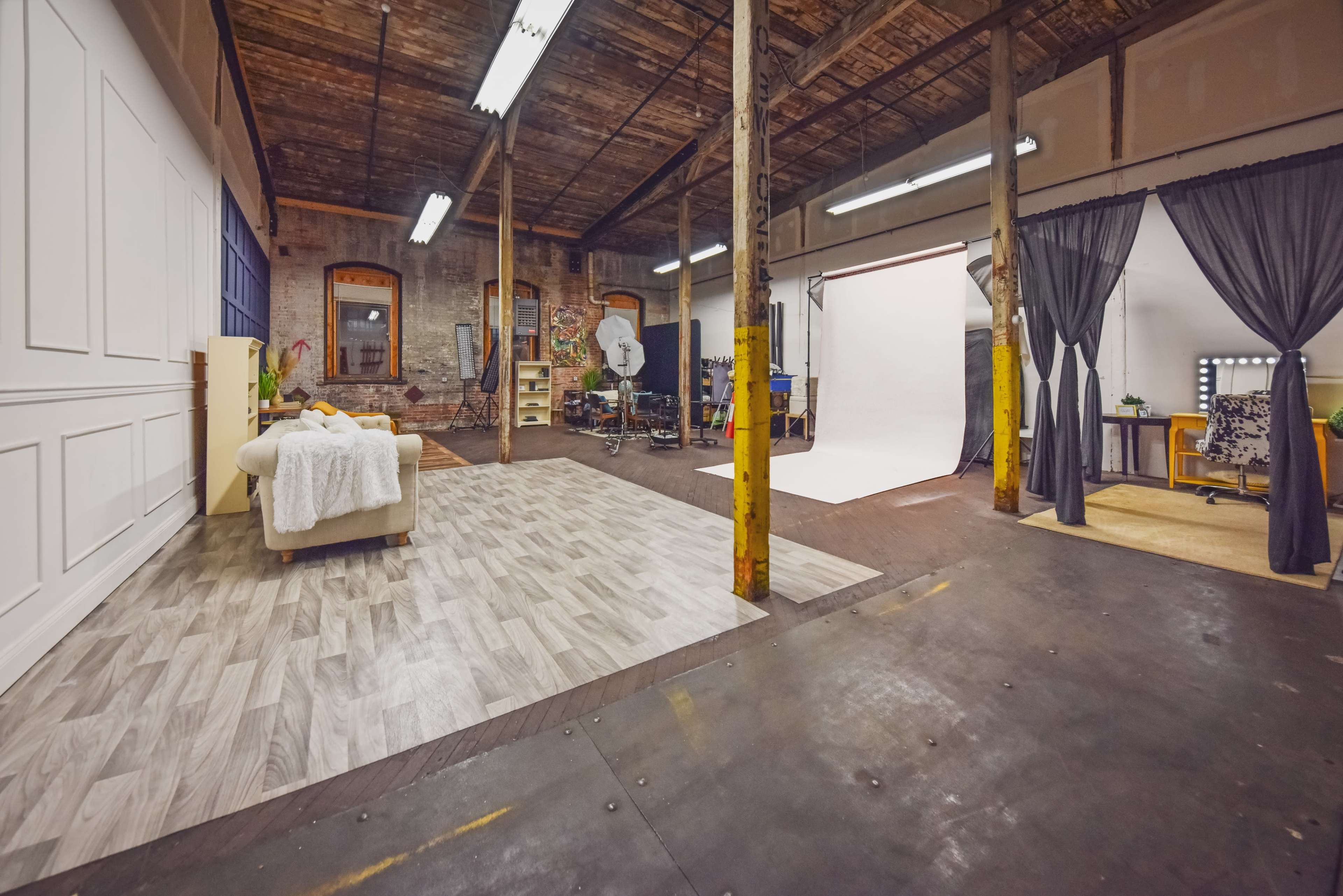 The image shows a spacious photography studio featuring a partially furnished area with a couch, a backdrop setup, and various equipment arranged along the walls.