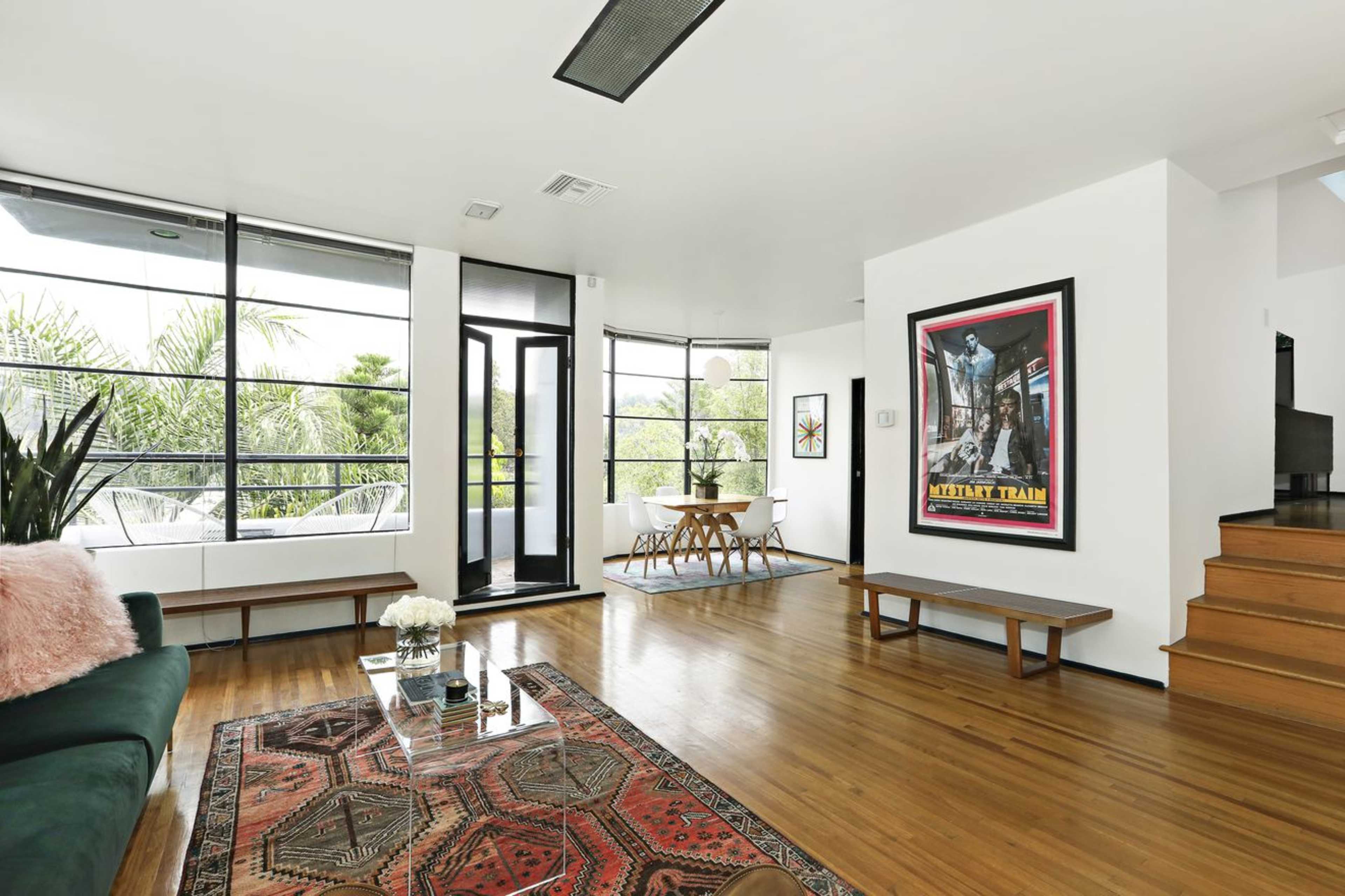 The image shows a modern living room featuring large windows, hardwood floors, a patterned area rug, and a dining table near a sliding glass door.