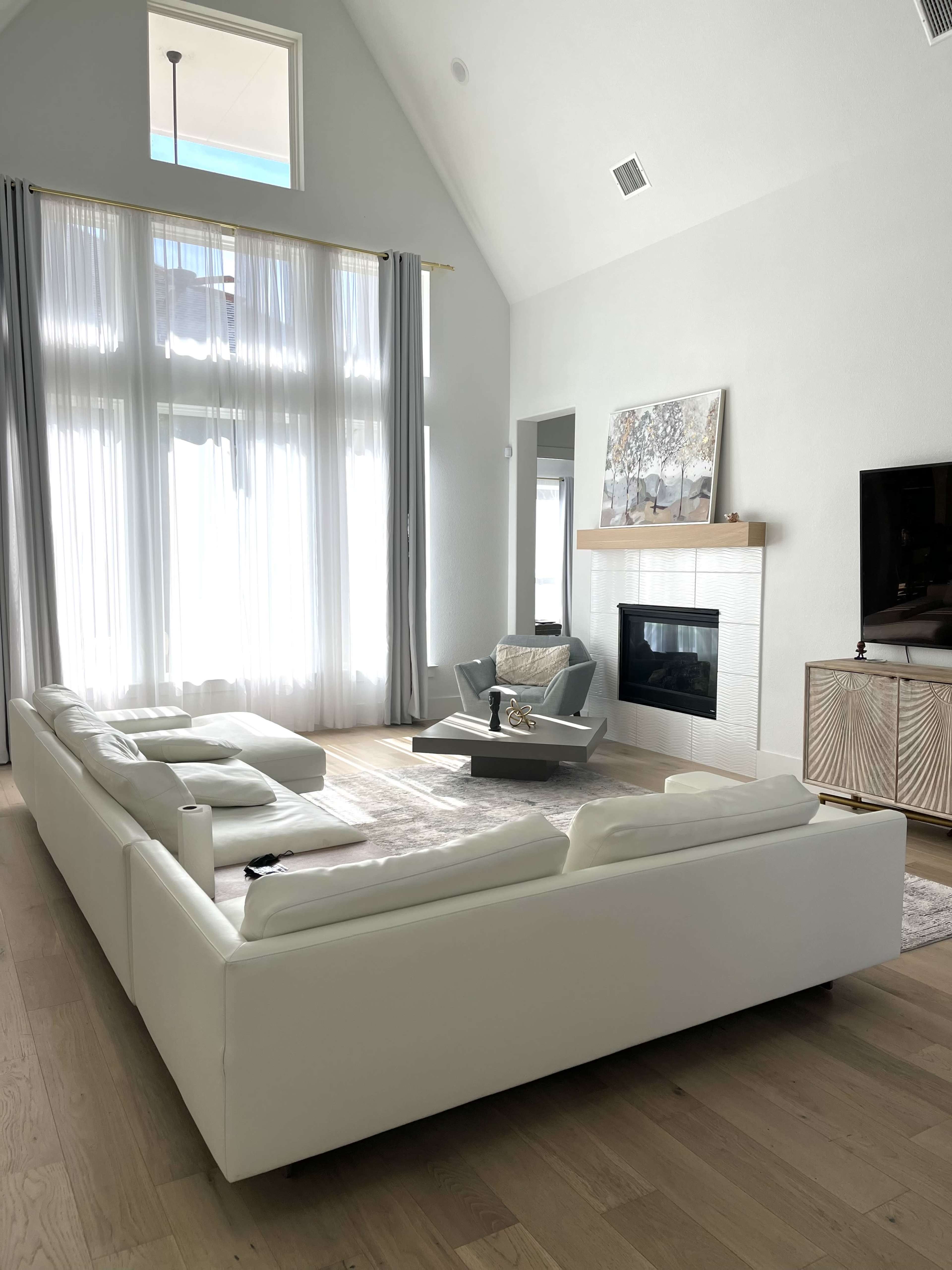 The image shows a modern living room with a white sectional sofa, a fireplace, a large window with sheer curtains, and a minimalist coffee table.