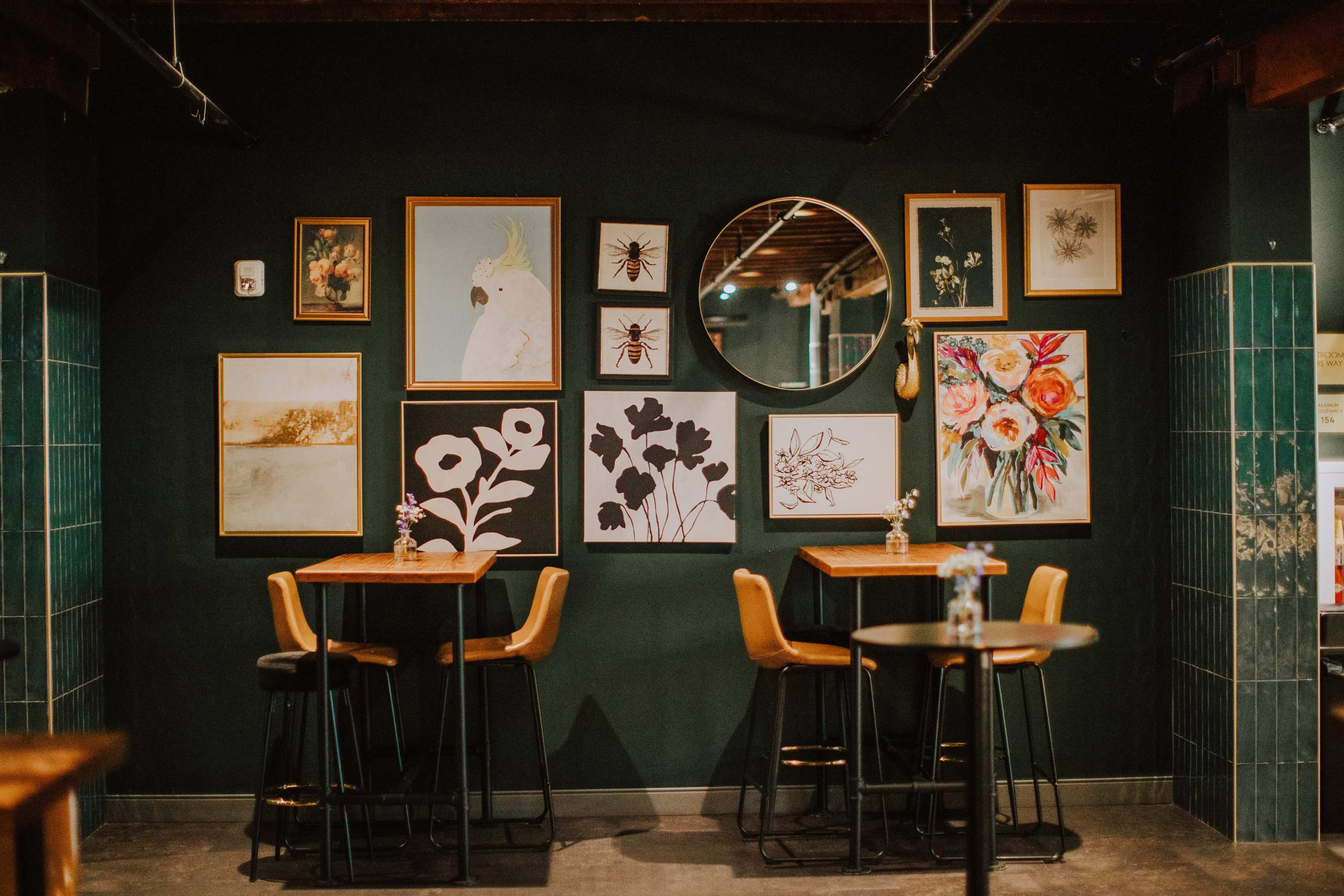 The image shows a cozy cafe interior featuring wooden tables and stylish chairs, with a gallery wall displaying various framed artworks and a round mirror.