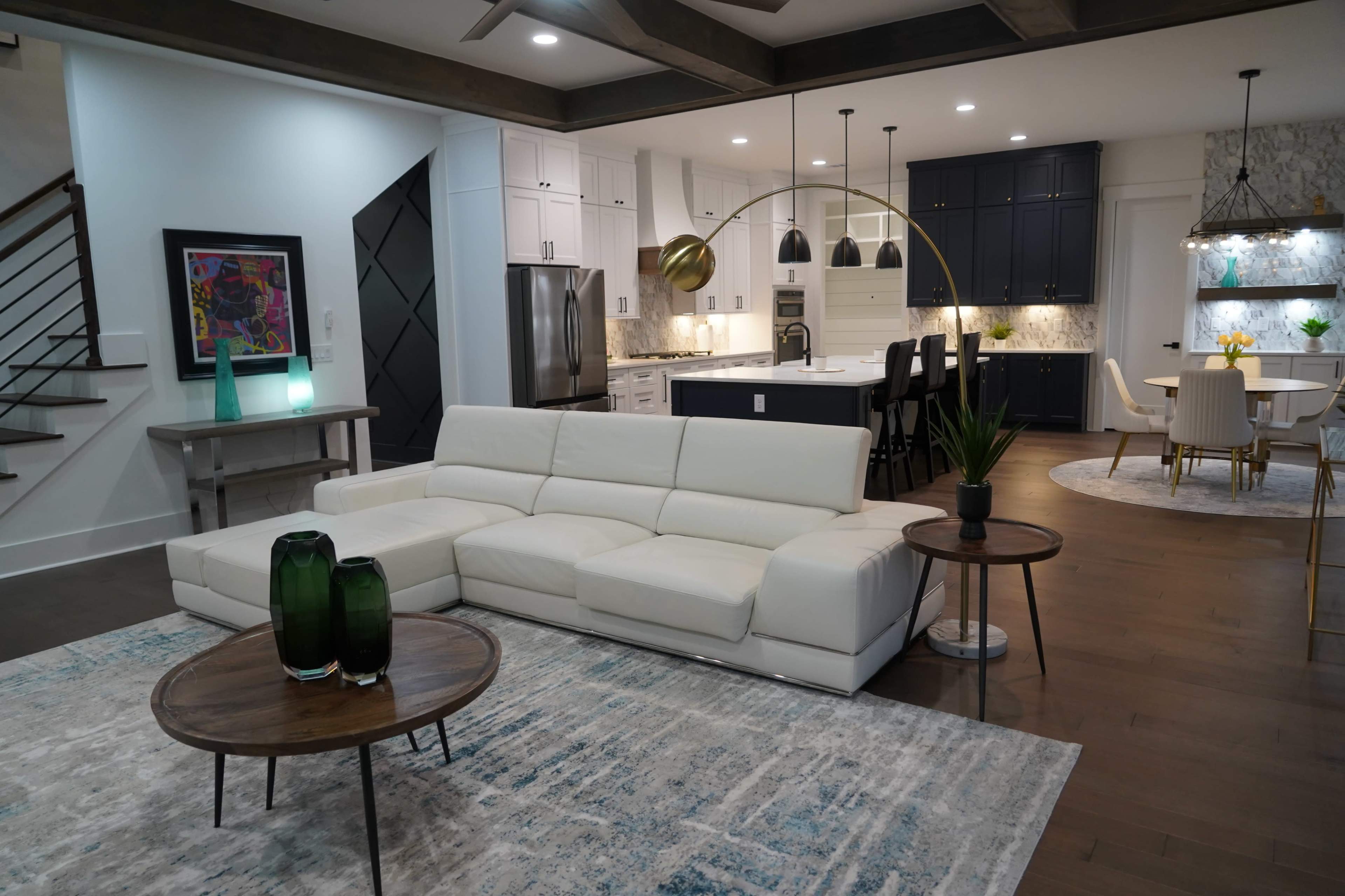 The image shows a modern living room featuring a white sectional sofa, two wooden side tables, and a kitchen area with dark cabinetry and marble countertops.