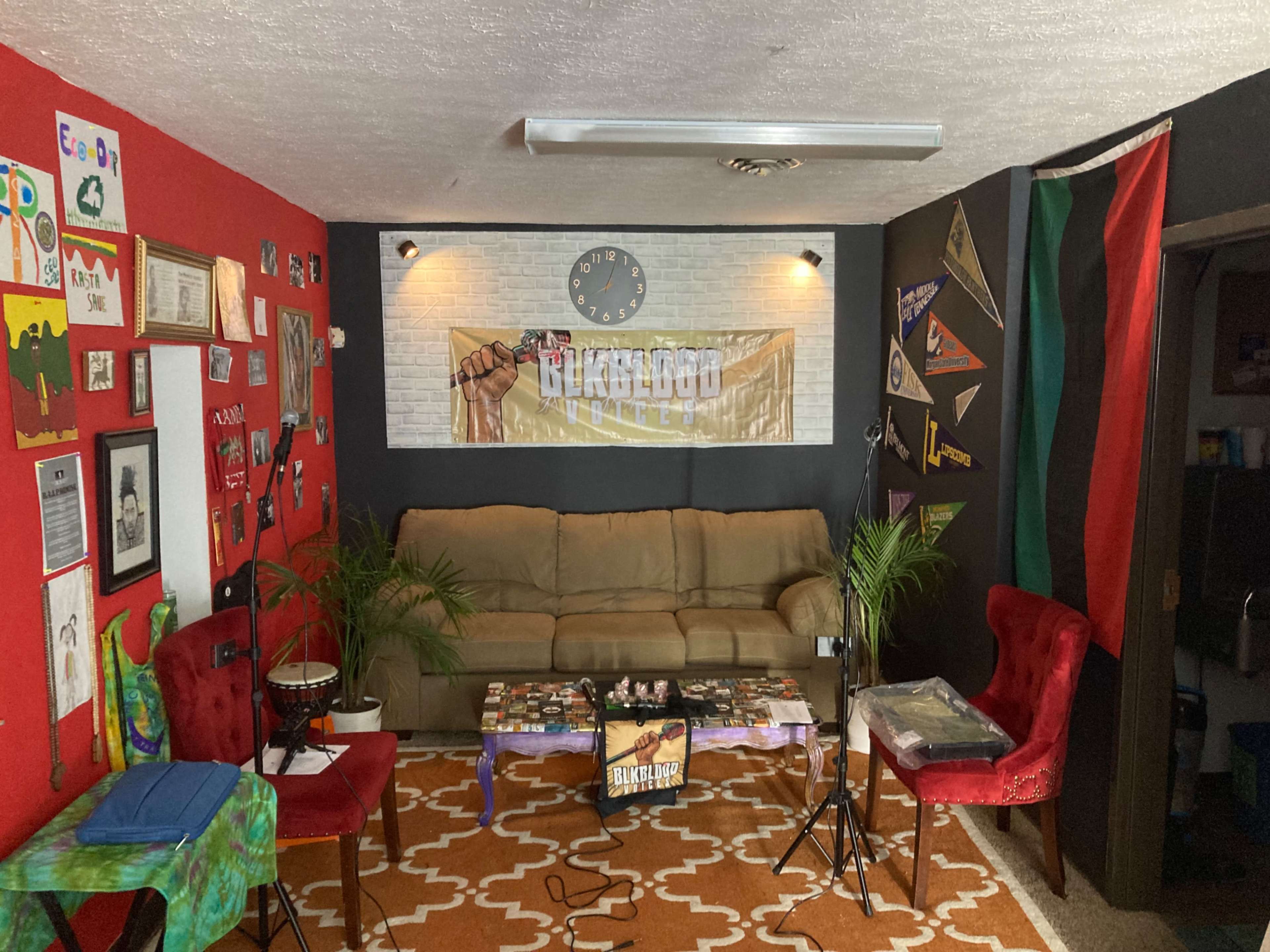 A cozy living room features a light brown couch, red and black walls adorned with various artworks and banners, a patterned rug, and two red chairs, with a table holding magazines in the center.