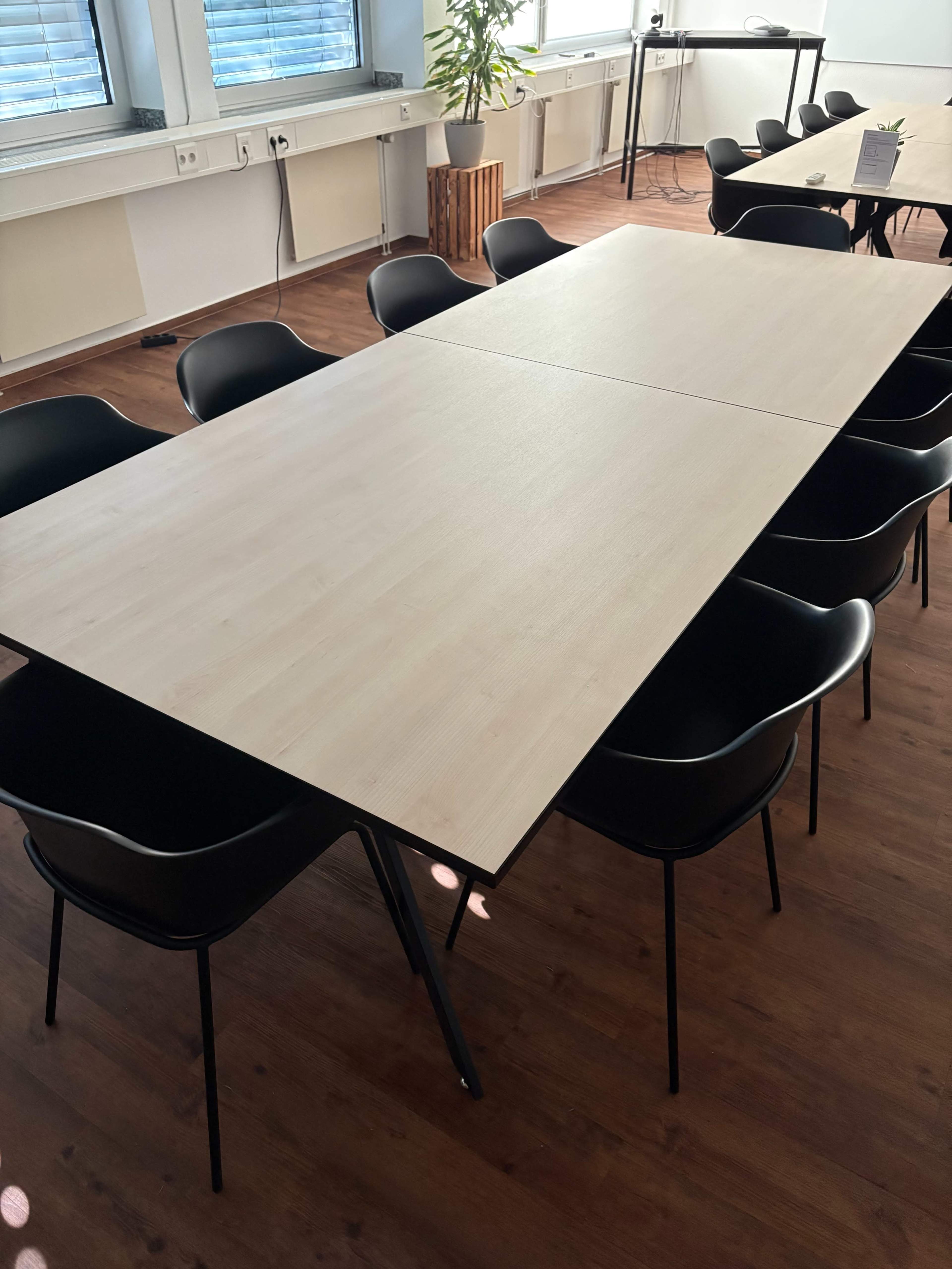 A large rectangular table with a light-colored surface is surrounded by black chairs in a well-lit room with wooden flooring.