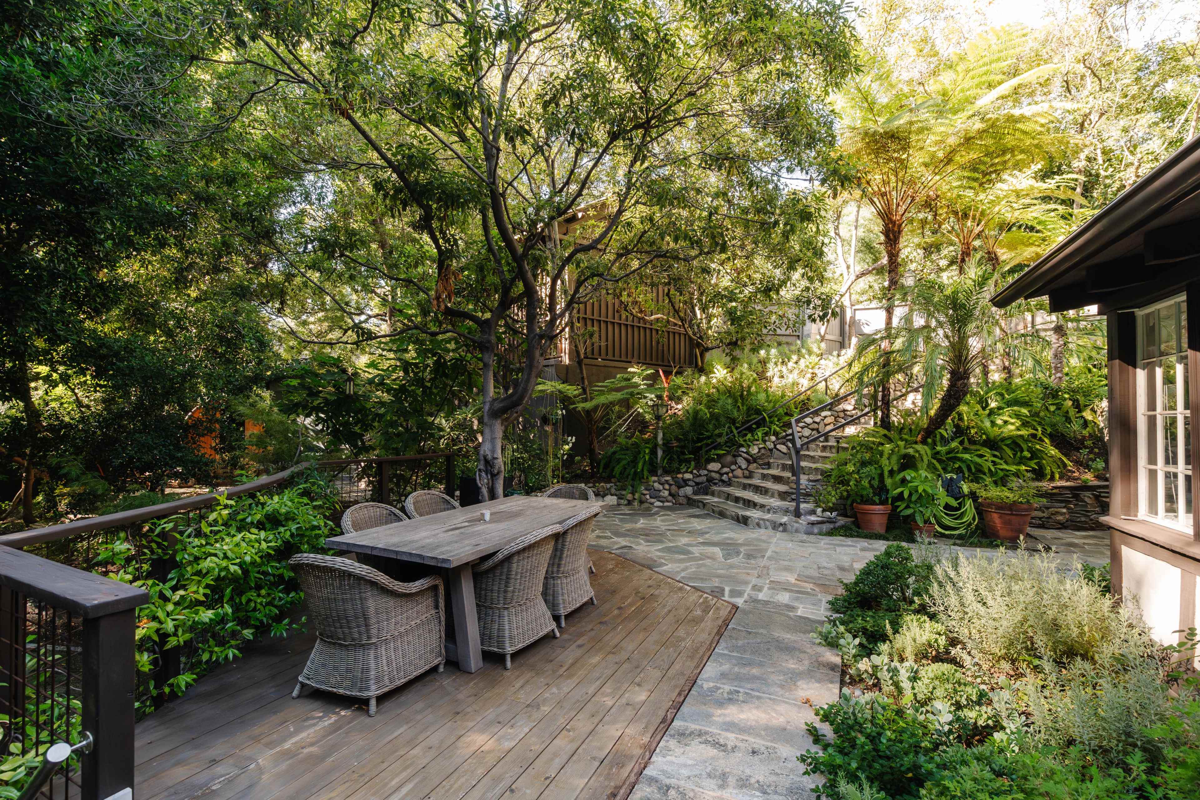 A wooden patio features a table surrounded by chairs, with a stone path and lush greenery leading to an elevated area in the background.
