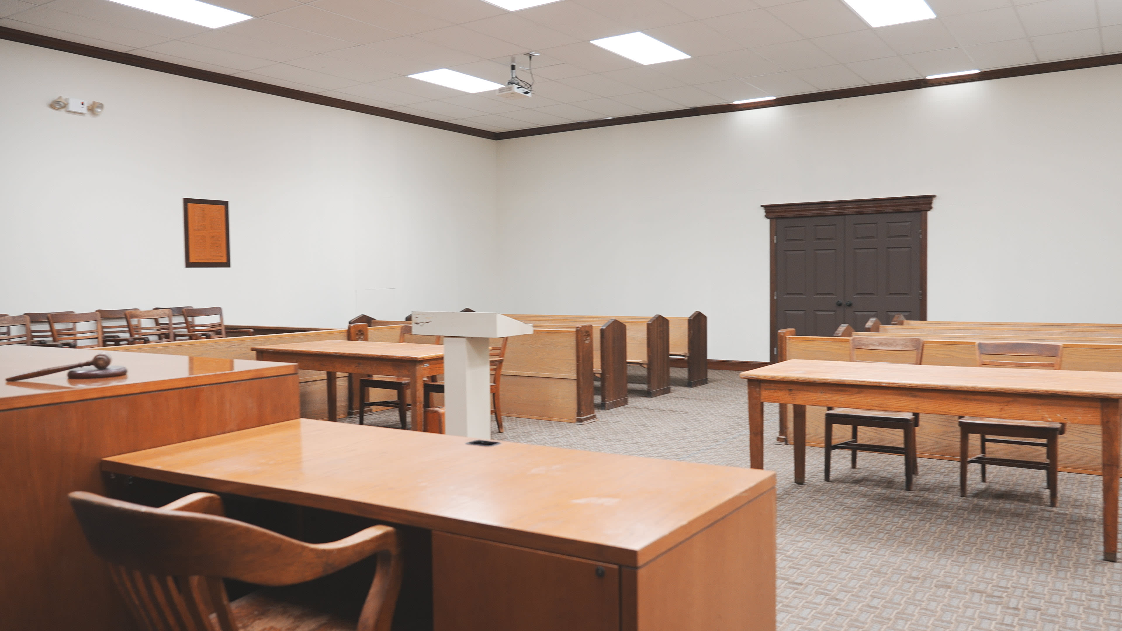 The image displays an empty courtroom with wooden tables, chairs, and a podium at the front.