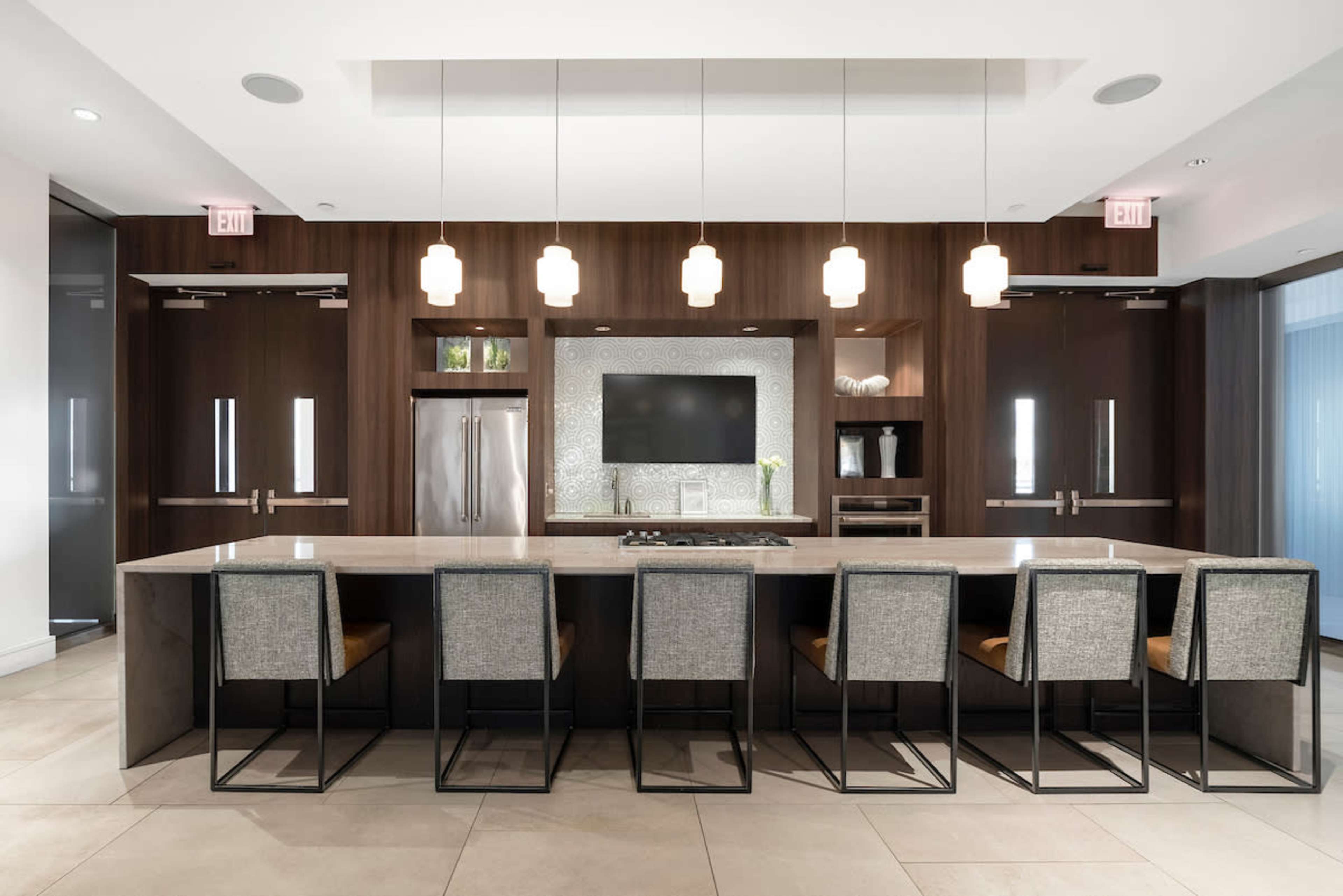 A modern kitchen features a long island with eight high-backed stools and a wall-mounted television above a sleek backsplash.