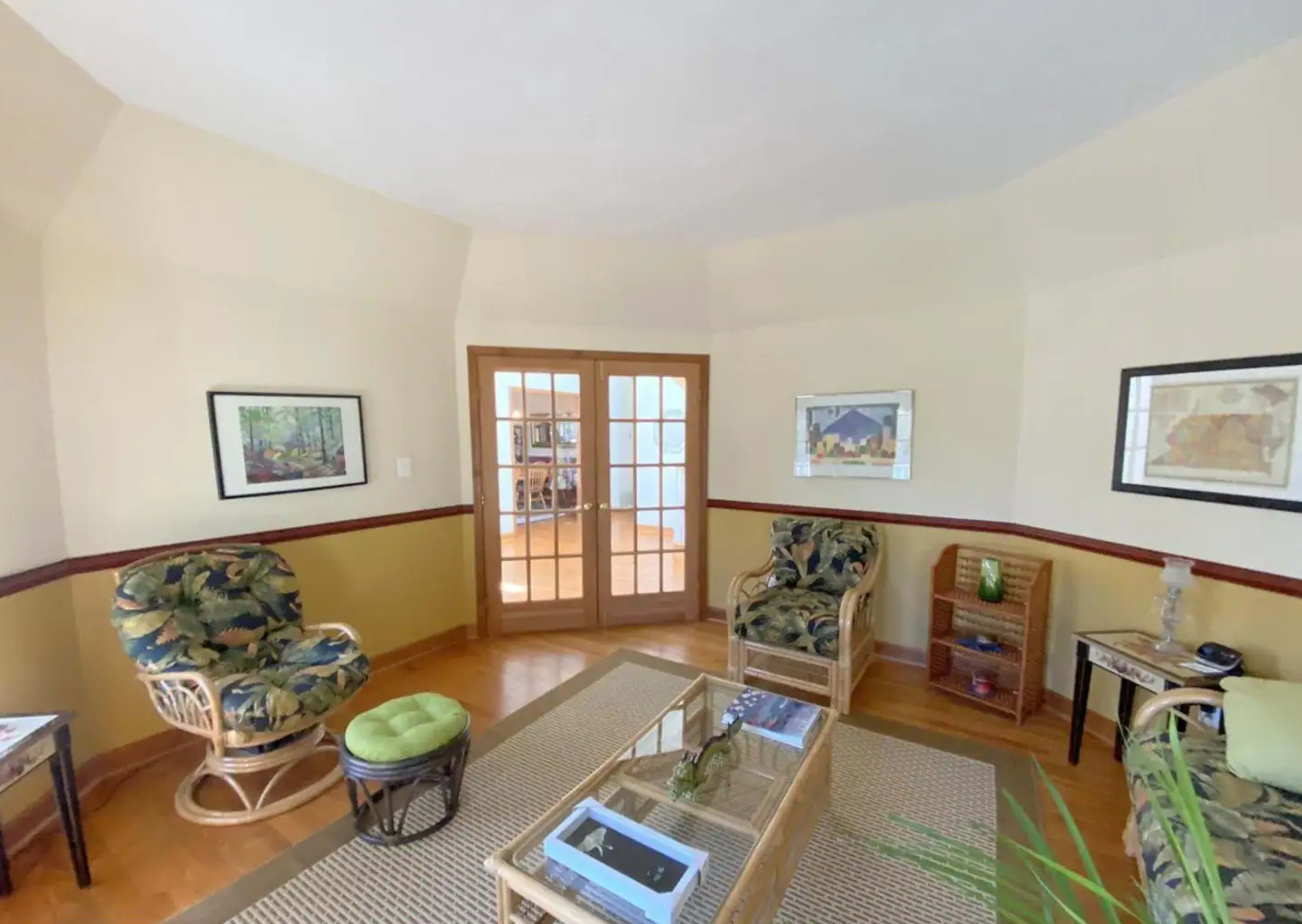 A cozy sitting area features two wicker chairs beside a glass-top coffee table, with artwork on the walls and a set of double doors leading to another room.