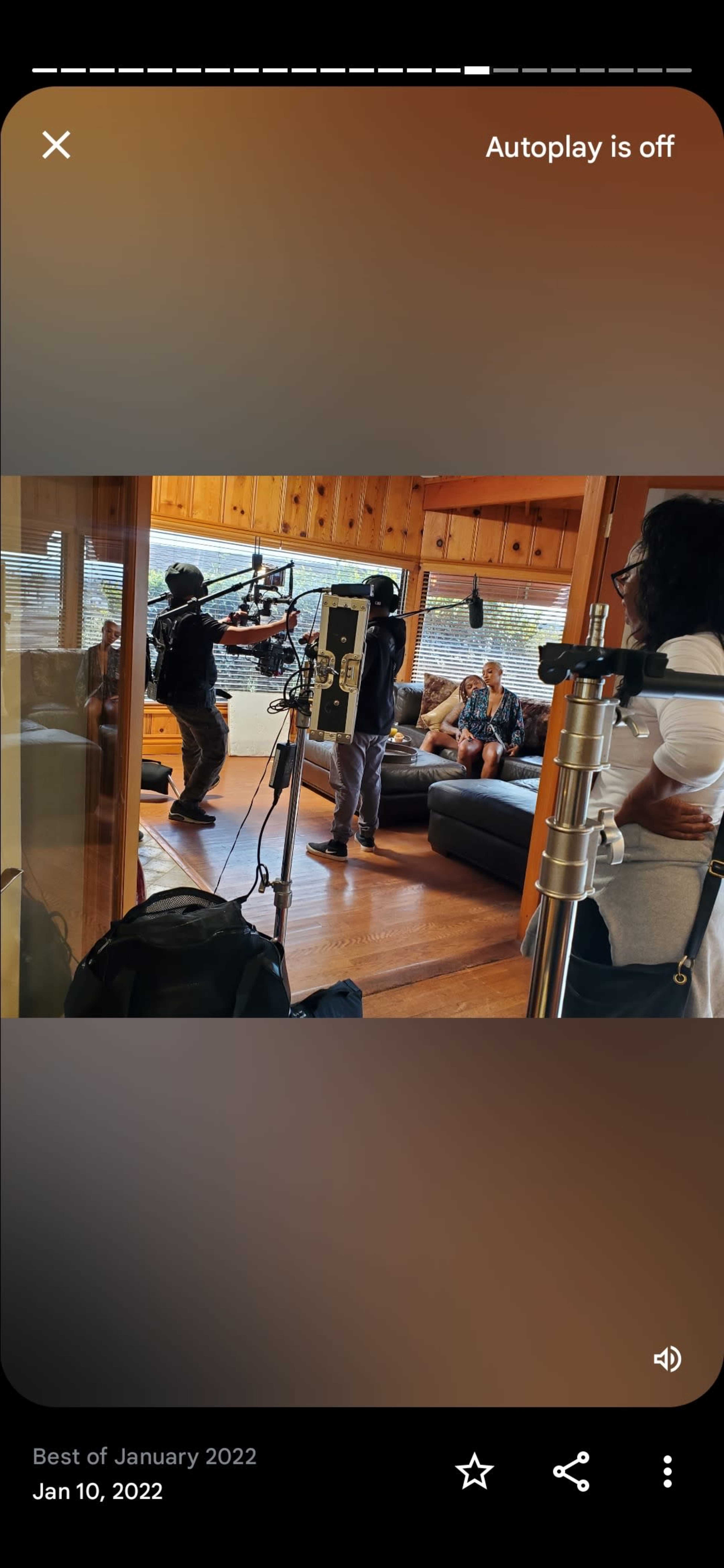 A film crew is set up in a wooden-paneled room, with a camera operator filming a person seated on a couch while another crew member prepares equipment.