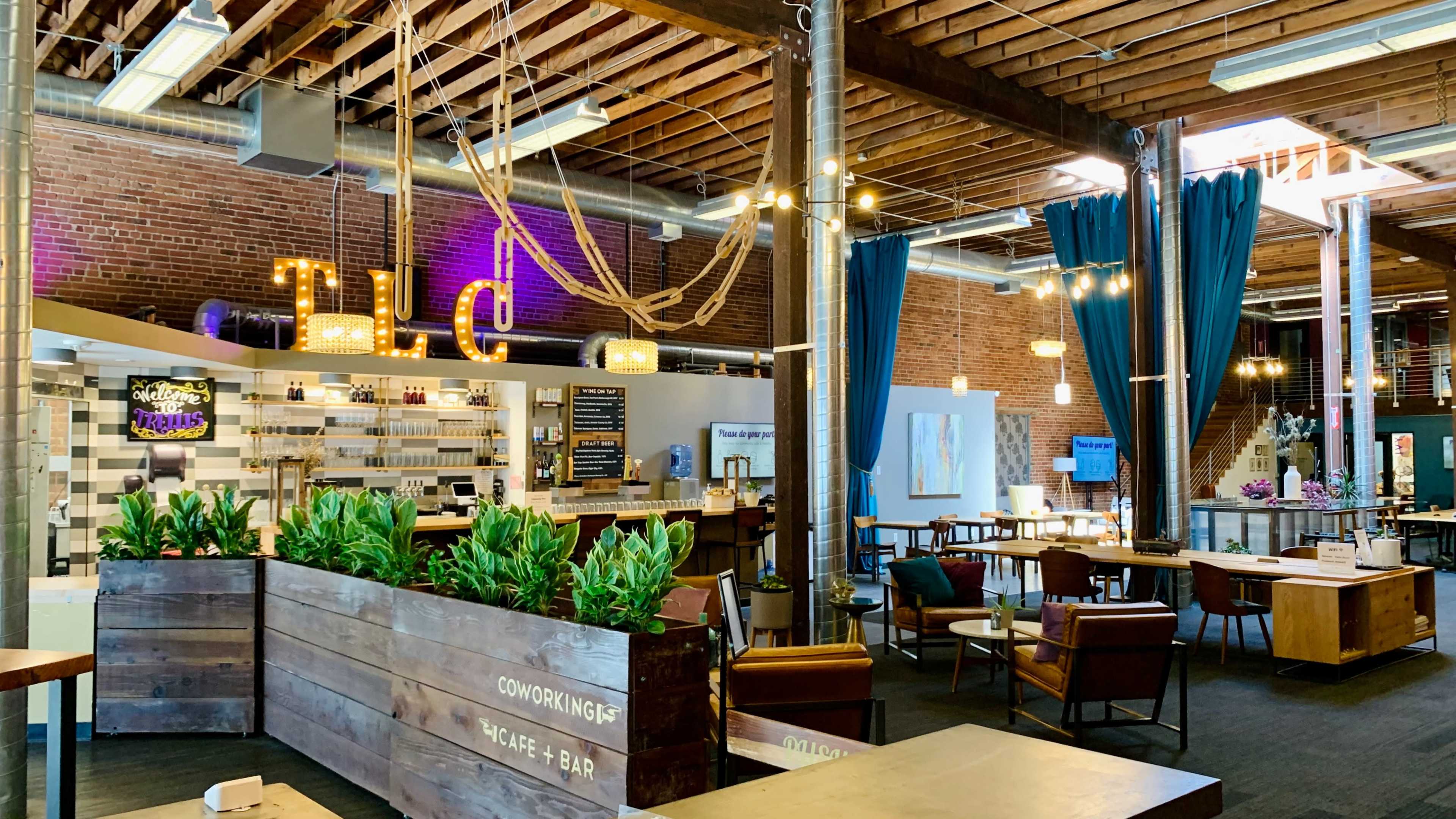 A spacious coworking space features wooden beams, a bar area, and various seating arrangements with plants and decorative lighting.