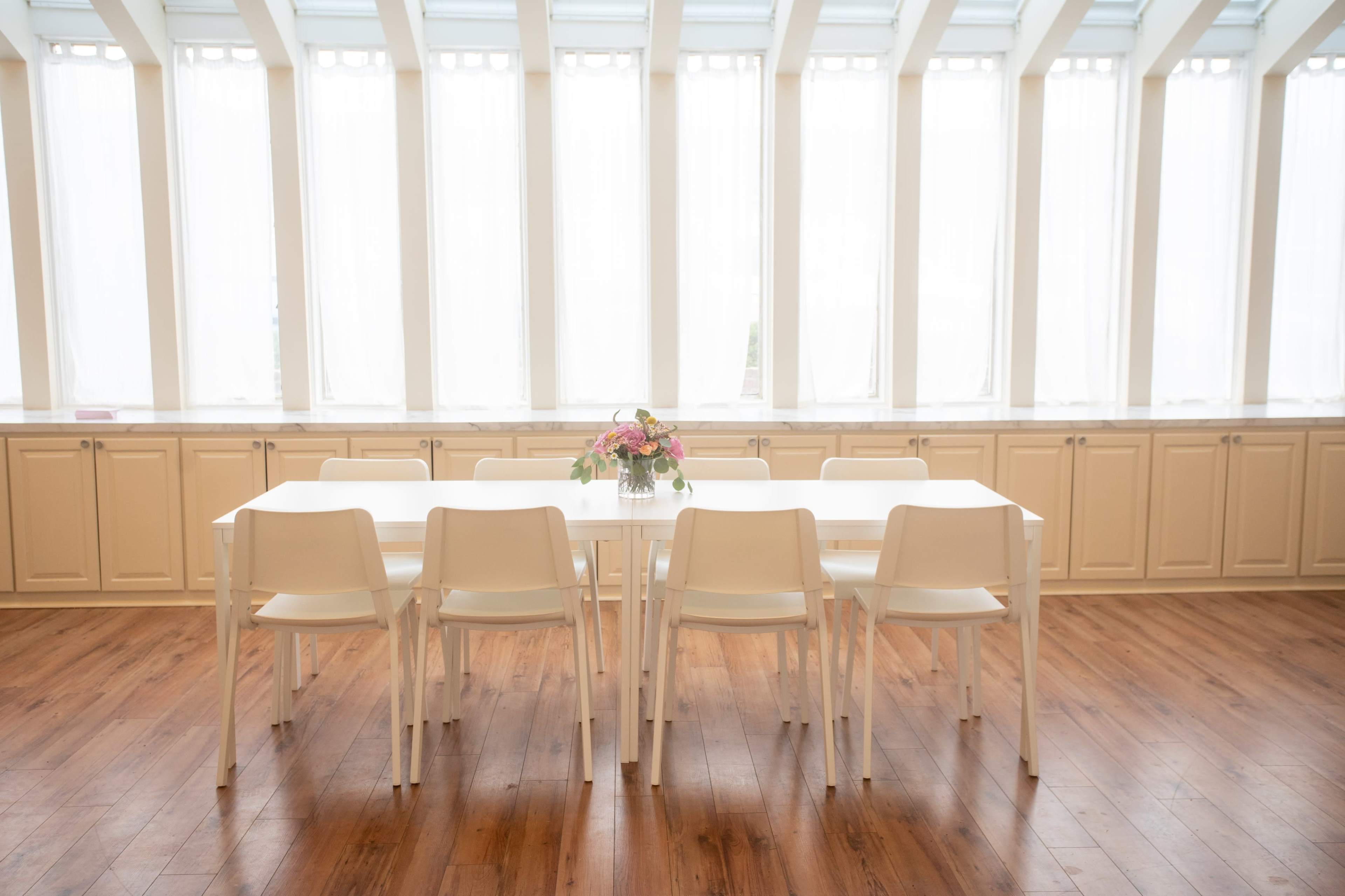 A long, white table with several white chairs is set in a room featuring large windows covered with sheer curtains, allowing natural light to fill the space.
