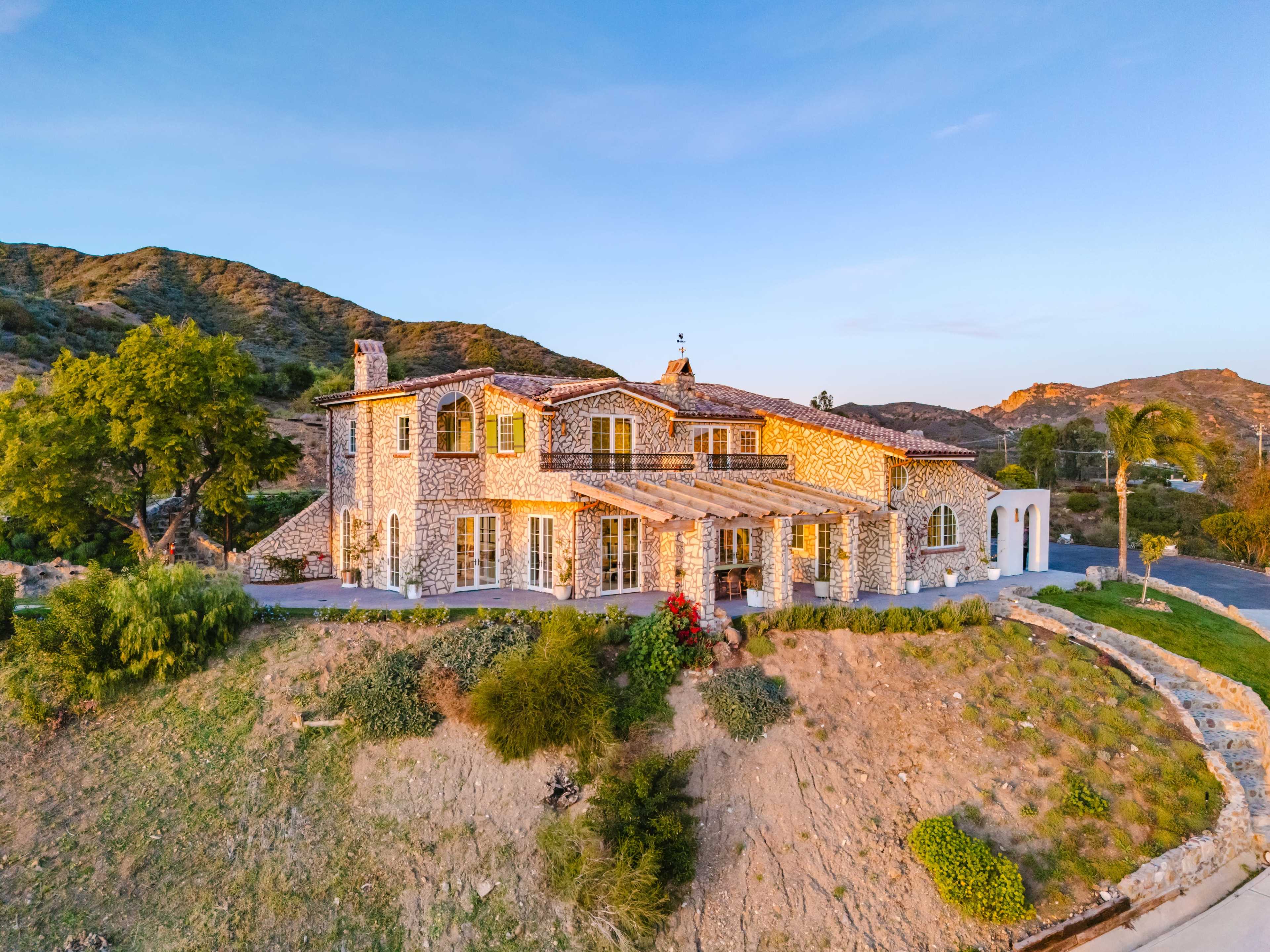 A large stone house with multiple windows and a tiled roof is situated on a hill, surrounded by greenery and mountains in the background.