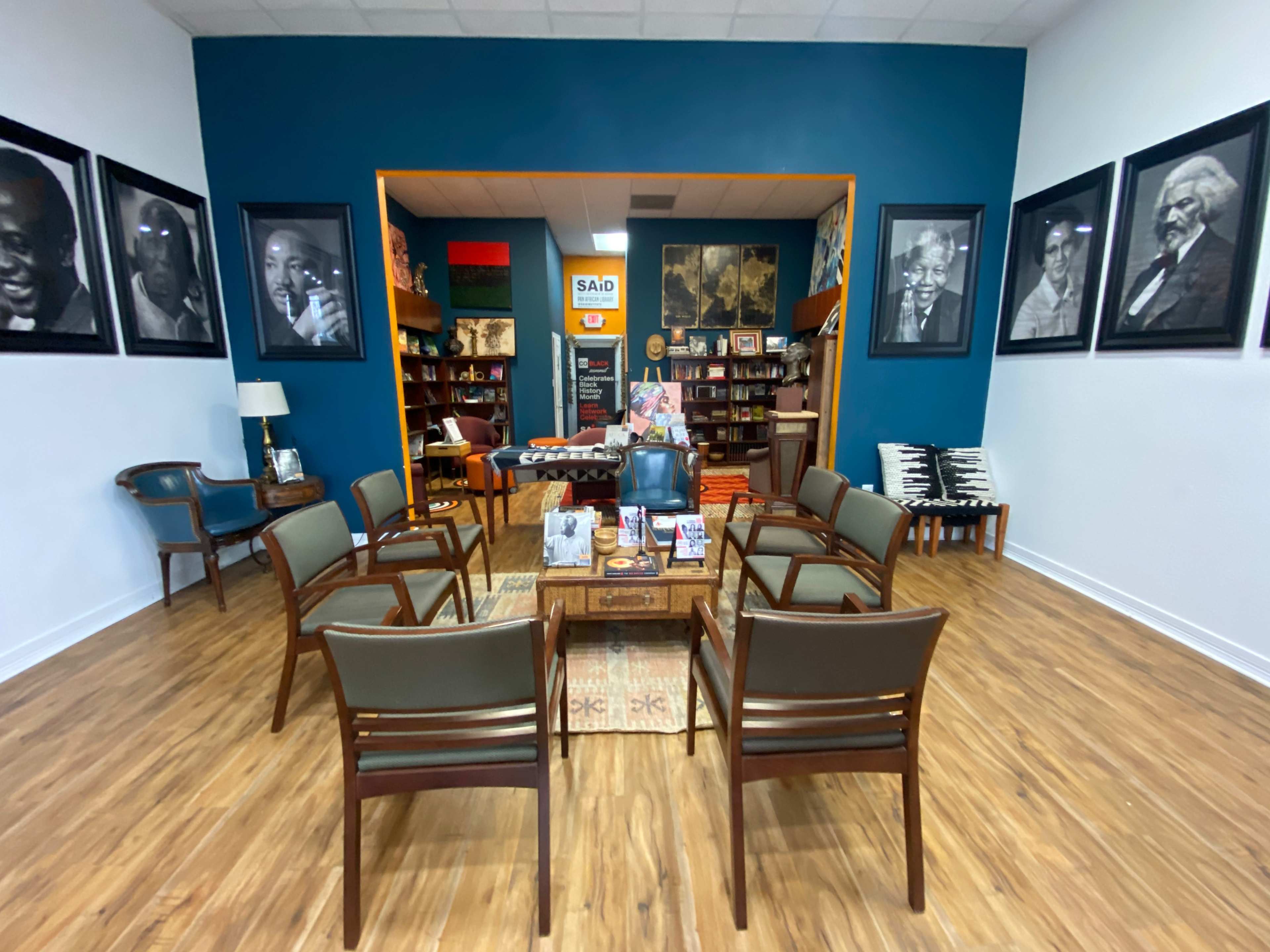 A spacious room with wooden flooring, featuring a circular arrangement of eight chairs around a wooden table, surrounded by large black-and-white portraits on the walls.