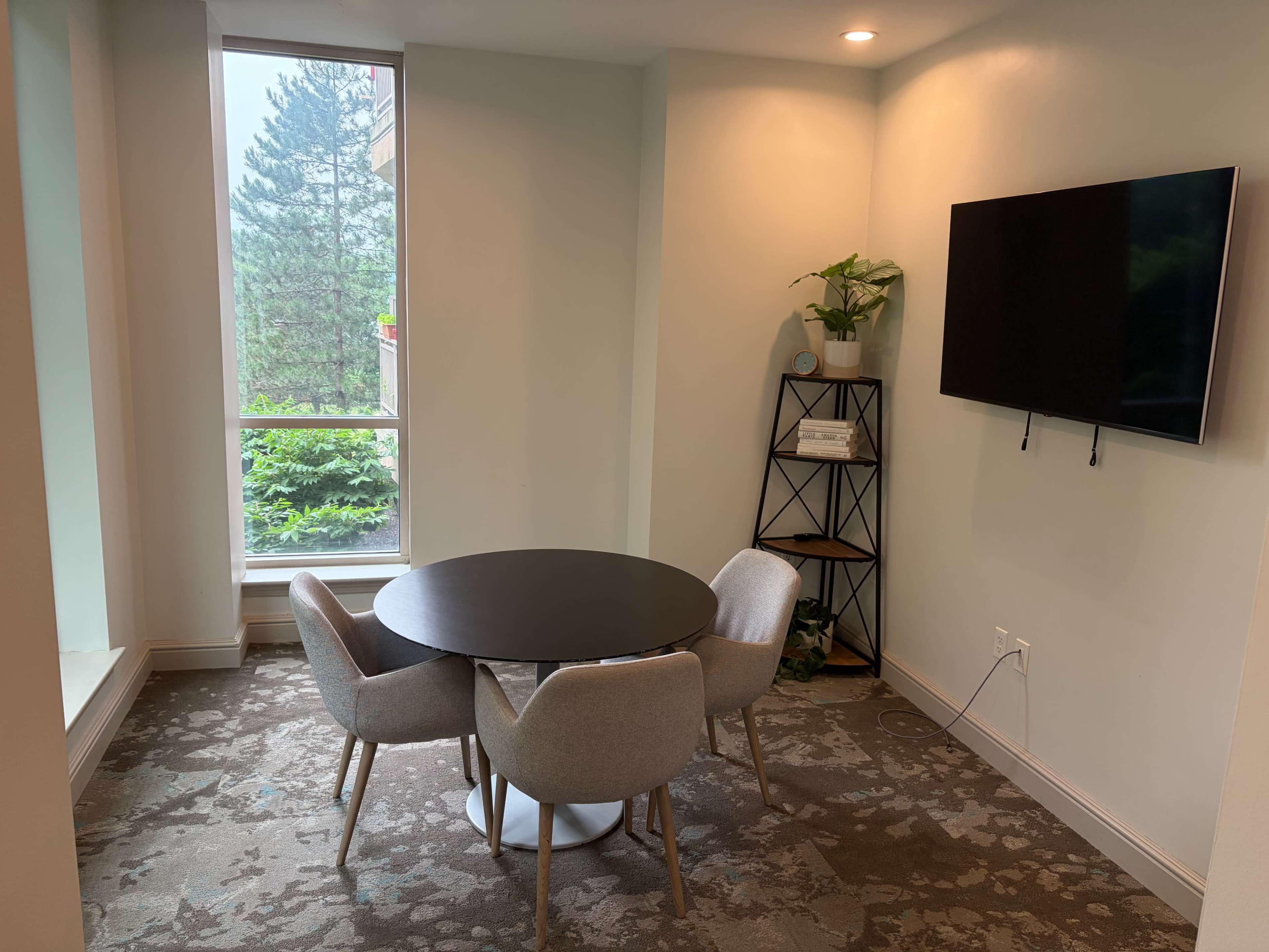 A small dining area features a round table surrounded by four upholstered chairs, with a shelf and a wall-mounted television nearby, and a large window overlooking greenery.
