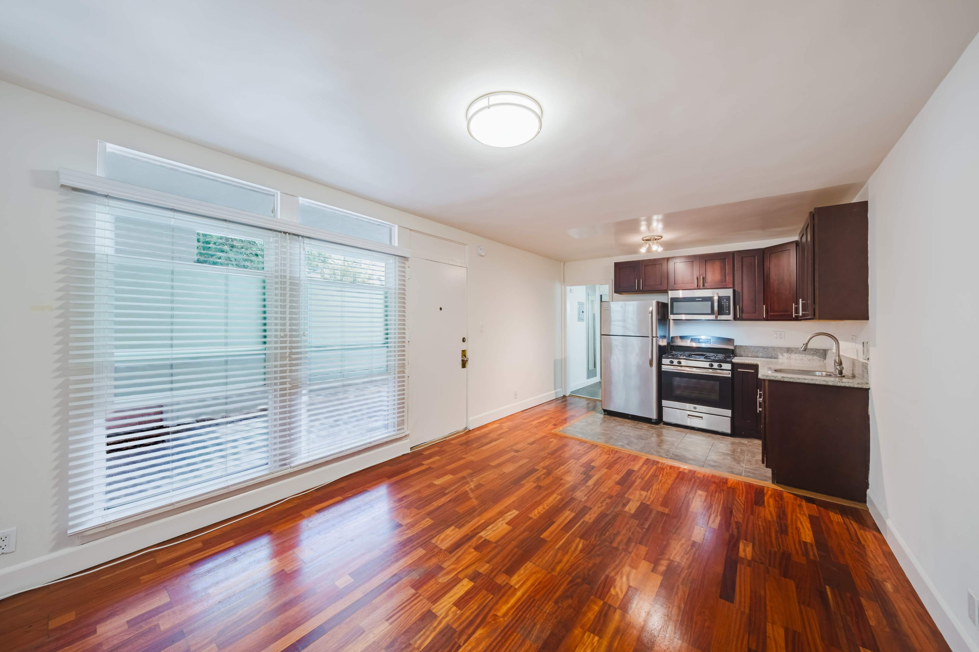 The image shows a modern kitchen and living space with hardwood floors, stainless steel appliances, and large windows.