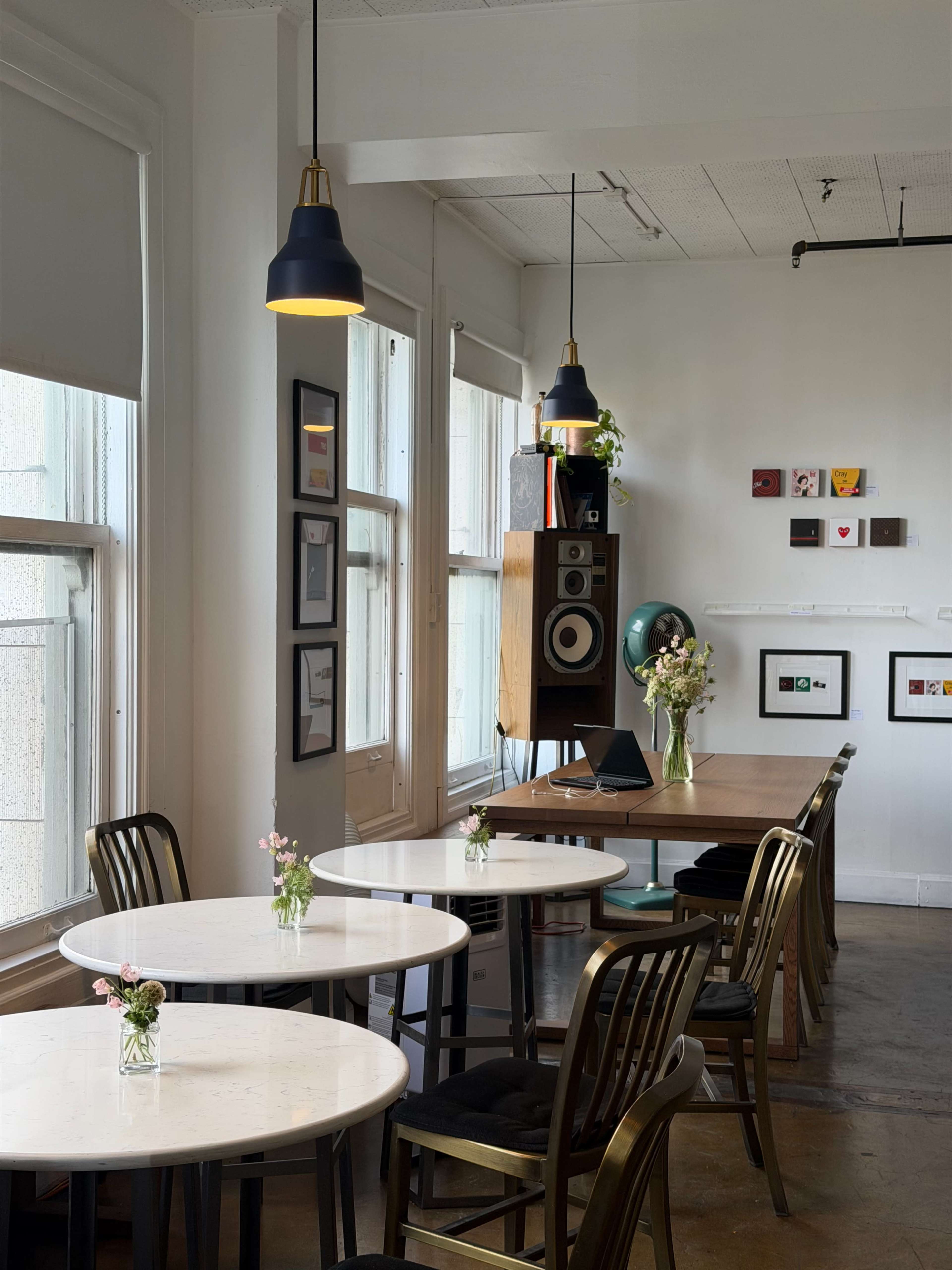 A brightly lit café features several tables with floral centerpieces, alongside a wall displaying a variety of framed artwork and a speaker system.